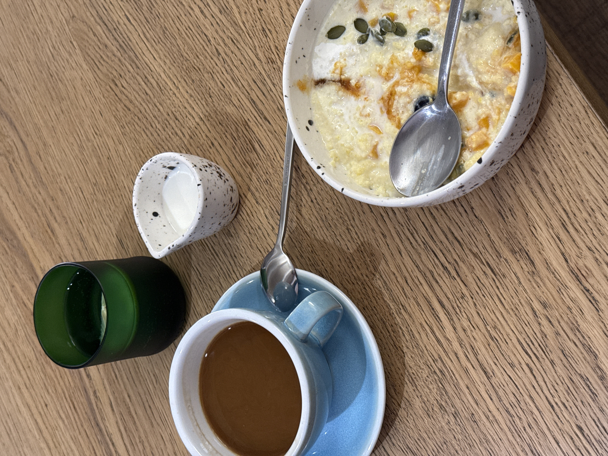 A high-angle shot captures a breakfast setting on a wooden table. The setting includes a blue teacup filled with coffee on a matching saucer, a speckled creamer containing milk, a green glass, and a speckled bowl filled with porridge, topped with pumpkin seeds and a spoon resting inside. Another spoon lies across the coffee cup. The image does not provide any information about people, actions, or interactions, nor does it confirm its location is in Chita, Russia.