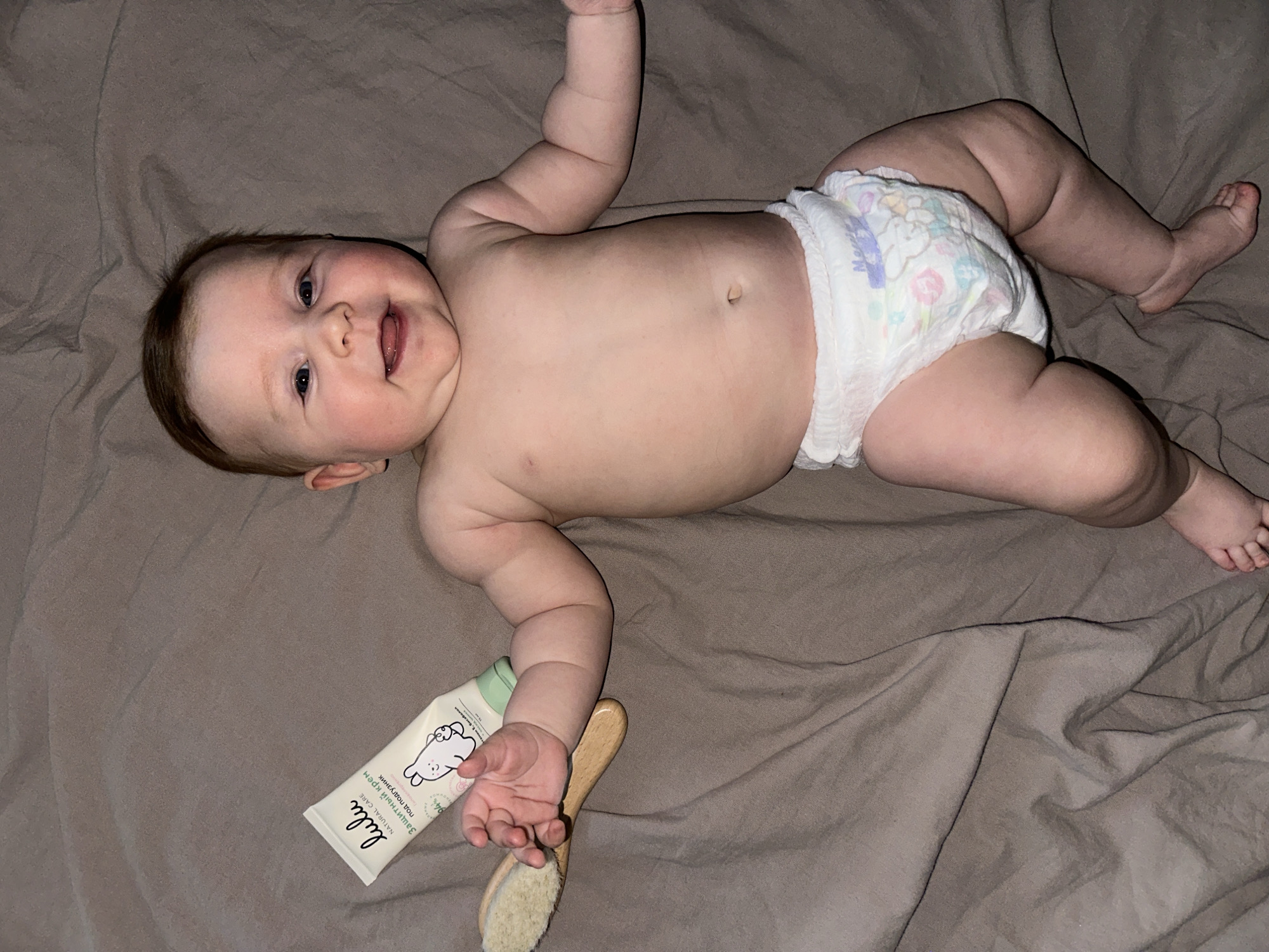 A baby is lying on a tan-colored fabric surface. The baby is on their back, wearing a white diaper with colorful patterns. The baby's arms are outstretched, and their face is visible with a smile. Near the baby's left hand is a white tube with a green cap, labeled "Lulu" with Russian text below, and a small wooden brush. The location is Chaikovsky, Russia.