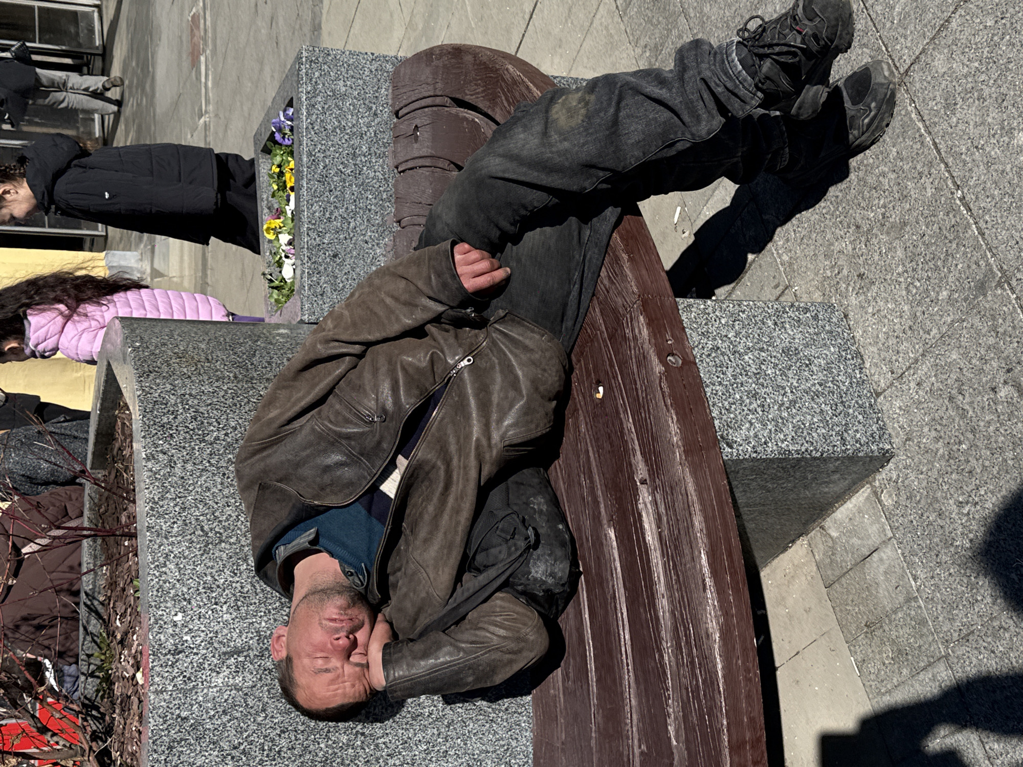The image shows a man lying on a park bench. He is wearing a brown leather jacket, blue sweater, and dark jeans with black shoes. The bench is made of dark wood with granite ends, including a planter with flowers. Other people are walking by in the background. The scene appears to be outdoors, on a sunny day.