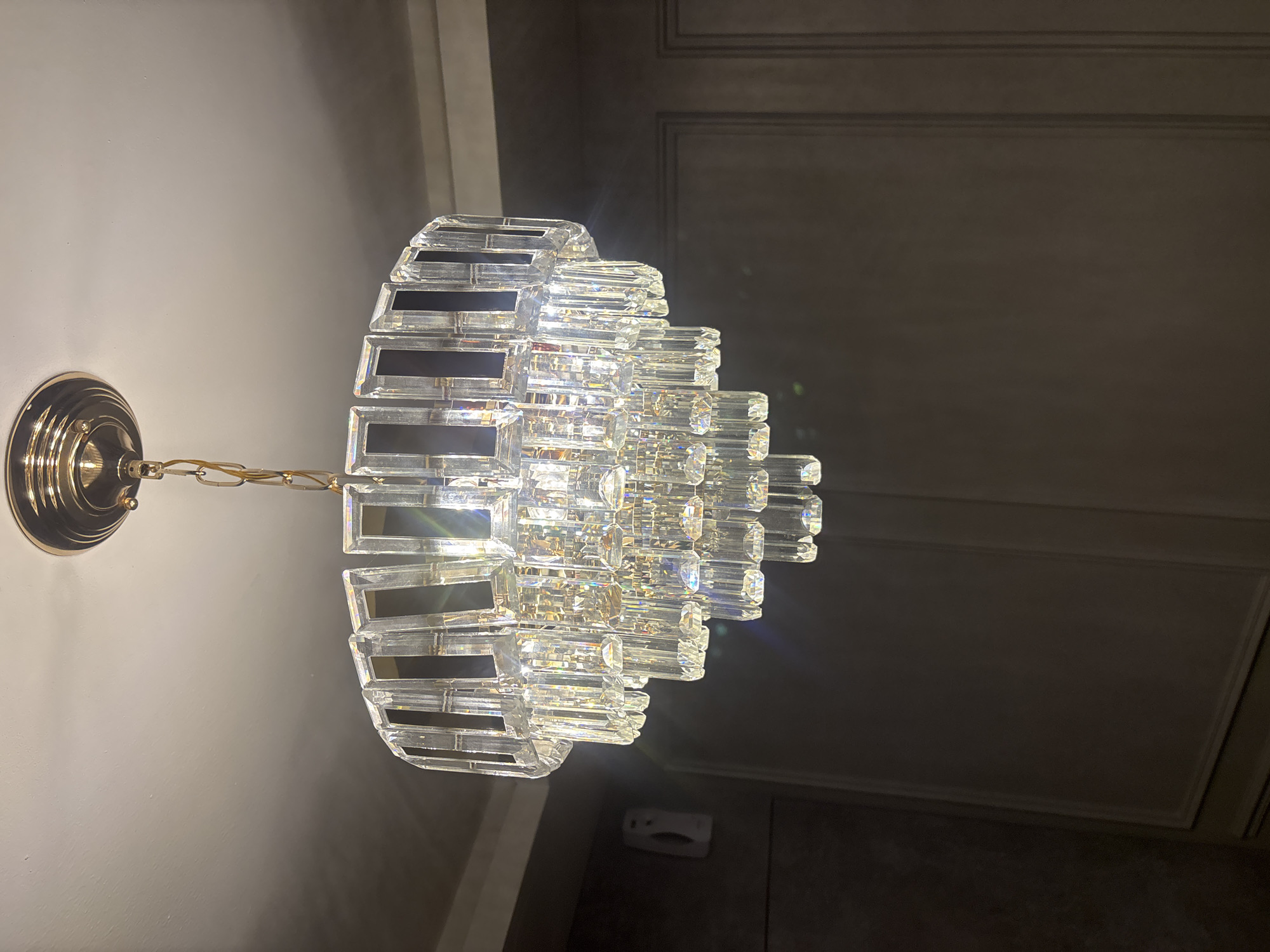A downward shot shows a tiered, crystal chandelier hanging from a gold ceiling mount by a gold chain. The chandelier features multiple layers of clear, rectangular crystal prisms. The ceiling is white, and the background consists of dark, paneled walls. To the left of the chandelier, a white wall-mounted device is visible. The scene appears to be indoors, in a room with dim lighting, likely in Pekanbaru, Indonesia.