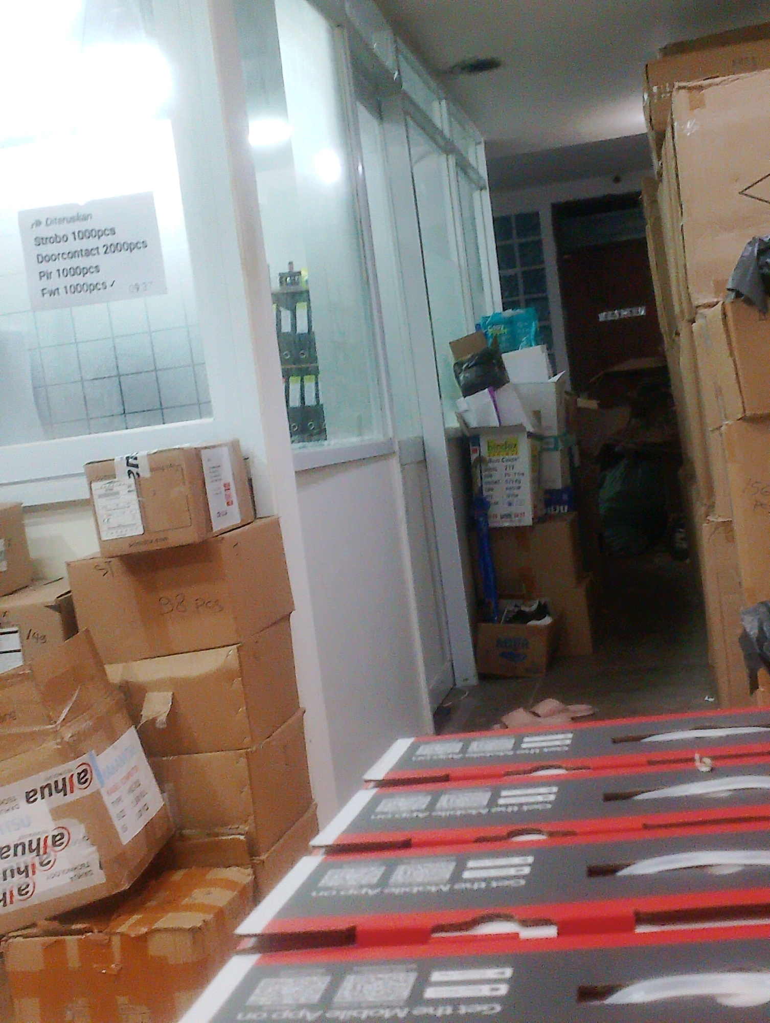 The image captures an interior scene that appears to be a warehouse or storage area. Boxes are stacked high, some made of cardboard and others appearing to be plastic containers. In the foreground, several flat, rectangular items with red and gray surfaces are lined up, each featuring a QR code and text that is partially visible but suggests a mobile application. To the left, a glass-paneled wall reveals another room or section of the building. A white sign with text in Indonesian is affixed to the glass, listing item quantities such as "Strobo 1000pcs" and "Doorcontact 2000pcs". The overall lighting suggests an indoor environment, possibly with some overhead illumination and natural light filtering through the glass. The image is focused on the storage and packaging elements within the space.