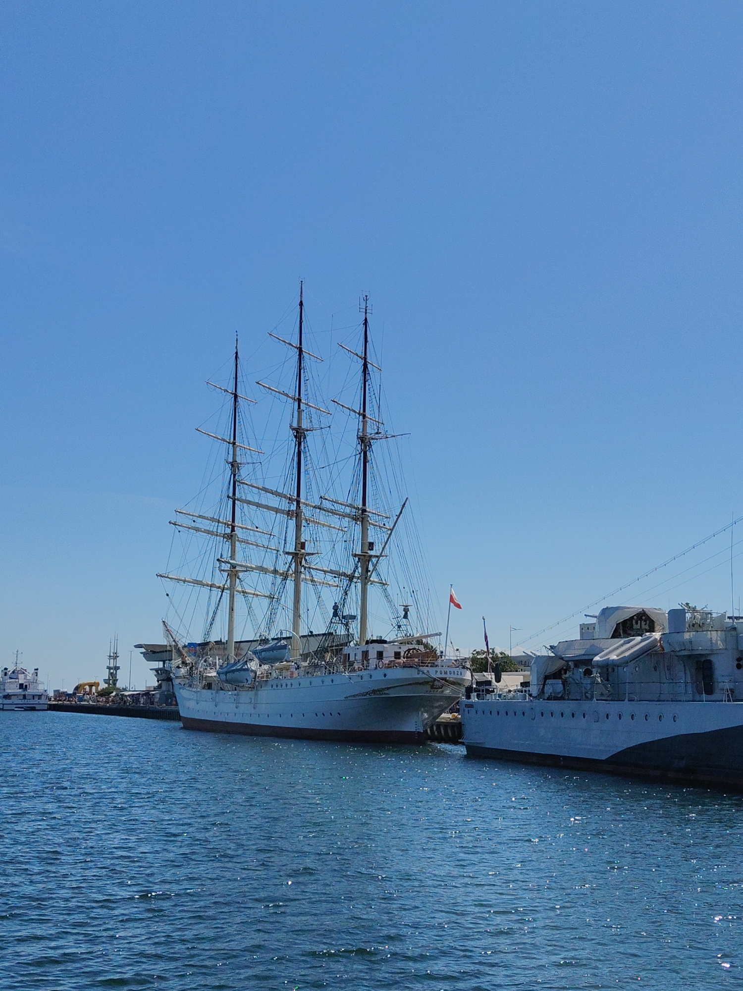 In Gdynia, Poland, on a clear day, a large sailing ship is docked in the port, its masts and rigging reaching towards the blue sky. To the right, a grey naval vessel with a distinct silhouette is also moored. The water in the harbor is a deep blue, reflecting the sunlight. In the background, other ships and port structures are visible, along with a small Polish flag flying from a flagpole on one of the vessels. The scene depicts a peaceful day at the maritime museum or a working port.