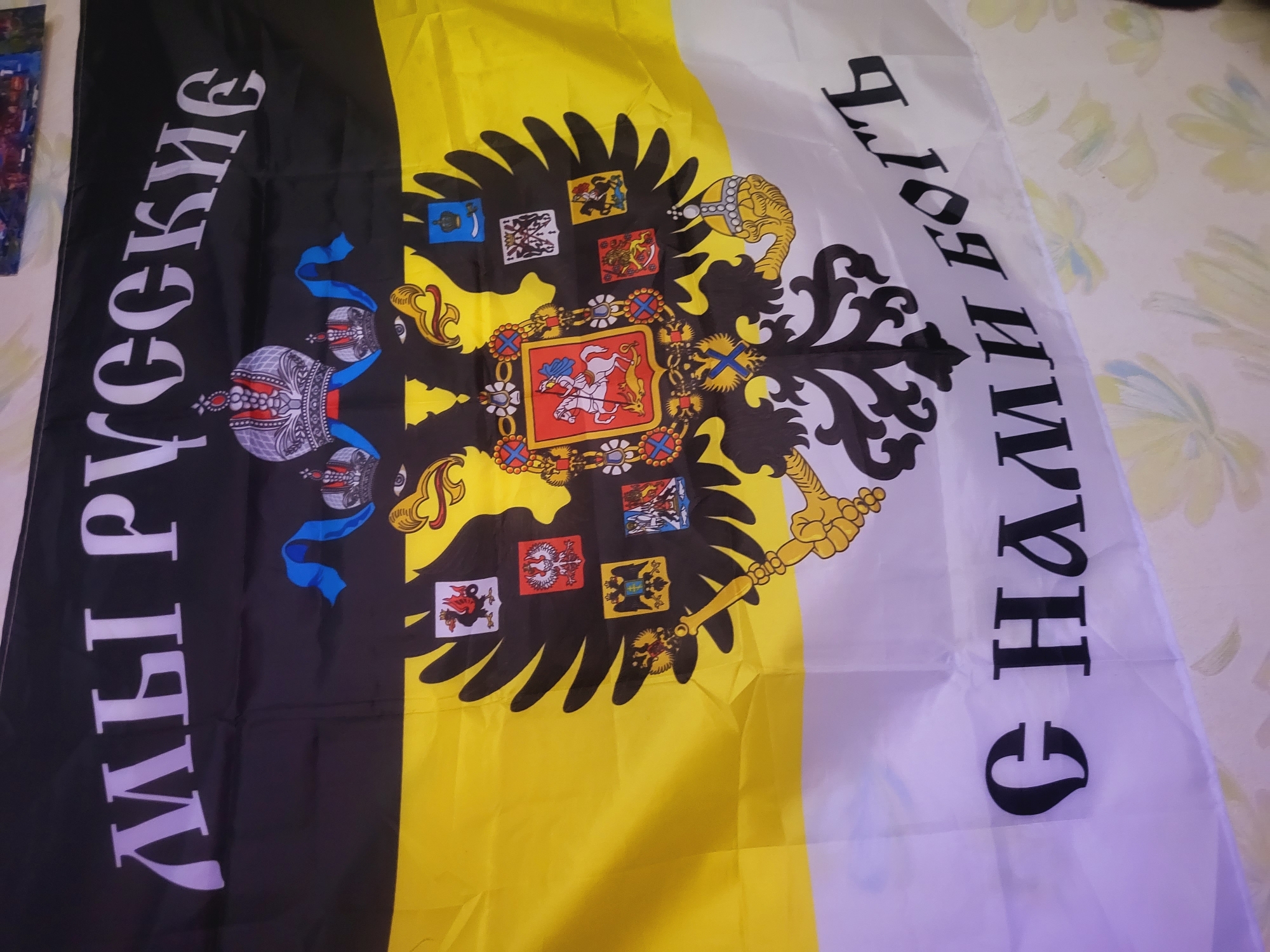 The image shows a flag with a black, yellow, and white tricolor design. On the black stripe to the left, vertical white text reads "МЫ РУССКИЕ" (We are Russian). In the center of the flag is a black double-headed eagle with a golden scepter and orb. The eagle's chest displays a shield with an image of Saint George slaying the dragon. Eight additional coats of arms are shown in circular frames within the eagle's wings. On the white stripe to the right, vertical black text reads "С НАМИ БОГЪ" (God is with us). The image provides no information about the location in the city of Зипуново, Russia.