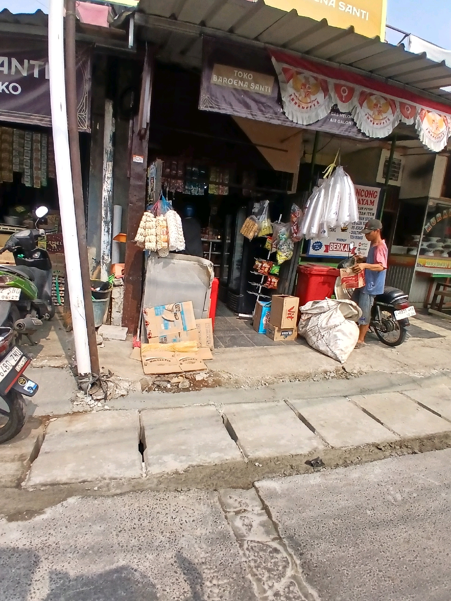 The image depicts a street-level view of a small shop in South Jakarta, Indonesia. In the foreground, a concrete sidewalk with visible expansion joints runs parallel to the street. Several concrete slabs are placed on the sidewalk, some of which appear to be new or under construction. To the left, the rear of a green and black motorcycle is visible, with a license plate reading "6480 ZMU 10-28". A white pole, likely part of the shop's structure or a utility pole, stands prominently on the sidewalk.
The shop itself is partially visible, with a dark awning overhead. A banner with the text "TOKO BAROENA SANTI" is displayed. Merchandise, including bags of goods and what appear to be snacks or household items, is hung outside the shop. Several cardboard boxes are stacked on the sidewalk, along with a large, light-colored woven bag. A red plastic bin is also present.
A man, wearing a blue vest over a pink shirt and denim shorts, stands near a black motorcycle parked in front of the shop. He is holding and examining a red and white item, possibly a product or receipt. He appears to be interacting with the goods or preparing them for transport.
The overall environment suggests a typical street scene in a commercial area, with elements of commerce, transportation, and urban infrastructure. The lighting indicates it is daytime, likely with strong sunlight.