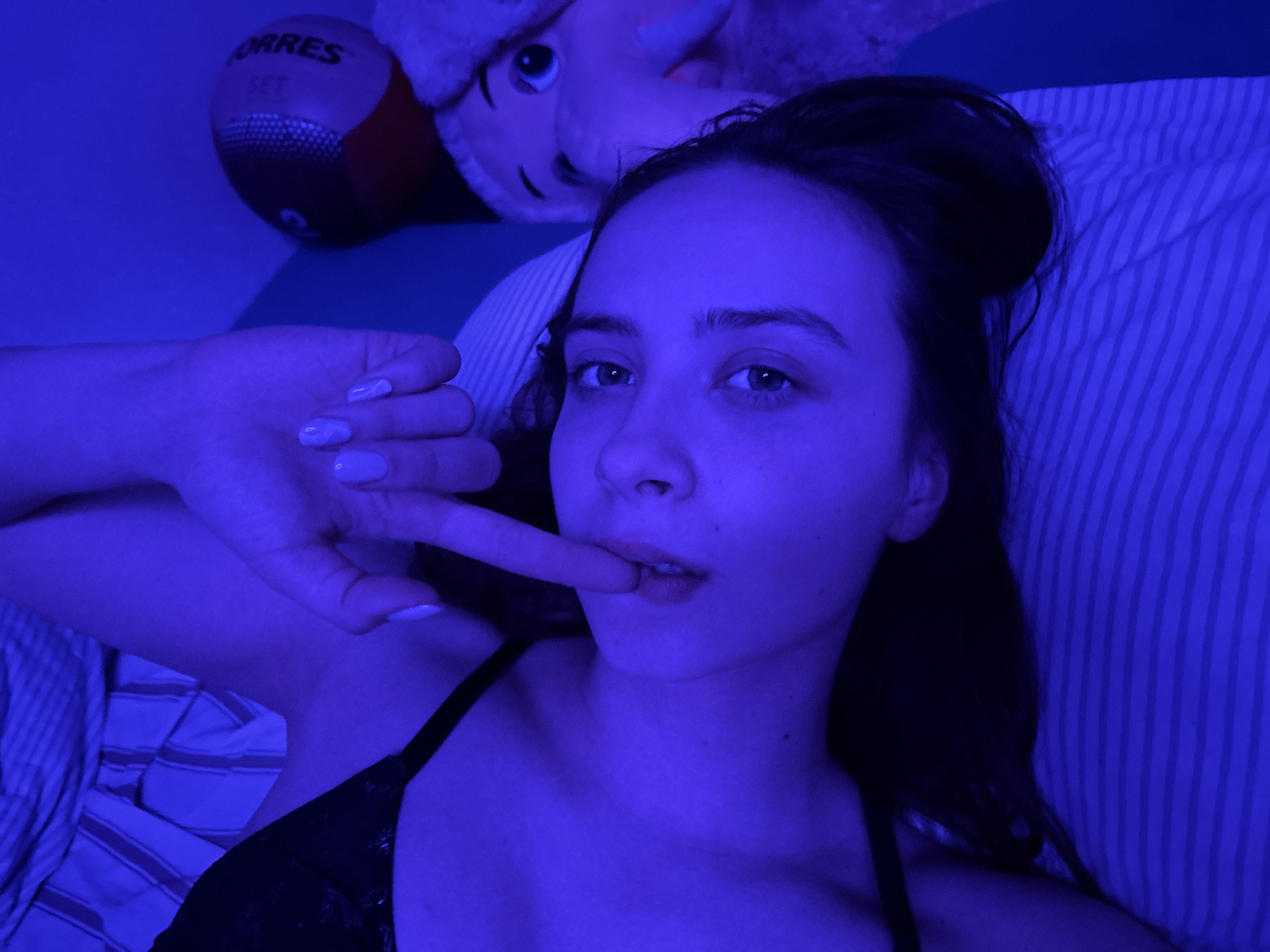 A young woman is captured in a close-up, high-angle shot, lying down indoors, likely on a bed, under pervasive blue ambient lighting. She looks directly into the camera with a neutral to slightly pensive expression. Her dark hair is styled in a bun. She wears a black top or bra. Her right hand is raised, with her index finger gently touching her lower lip. Her fingernails, painted a light color, appear purple due to the blue light.
Behind her head, the upper portion of a light-colored stuffed animal with prominent eyes is visible. To the left, a red and black medicine ball is partially seen, displaying the text "ORRES" and "SET." The pillow and bedding feature a striped pattern, appearing in shades of blue and muted grey under the strong blue illumination. The distinct blue light suggests an artificial light source, creating a moody ambiance, likely indicating an evening or nighttime setting. The scene is set in Прикамский, Russia.