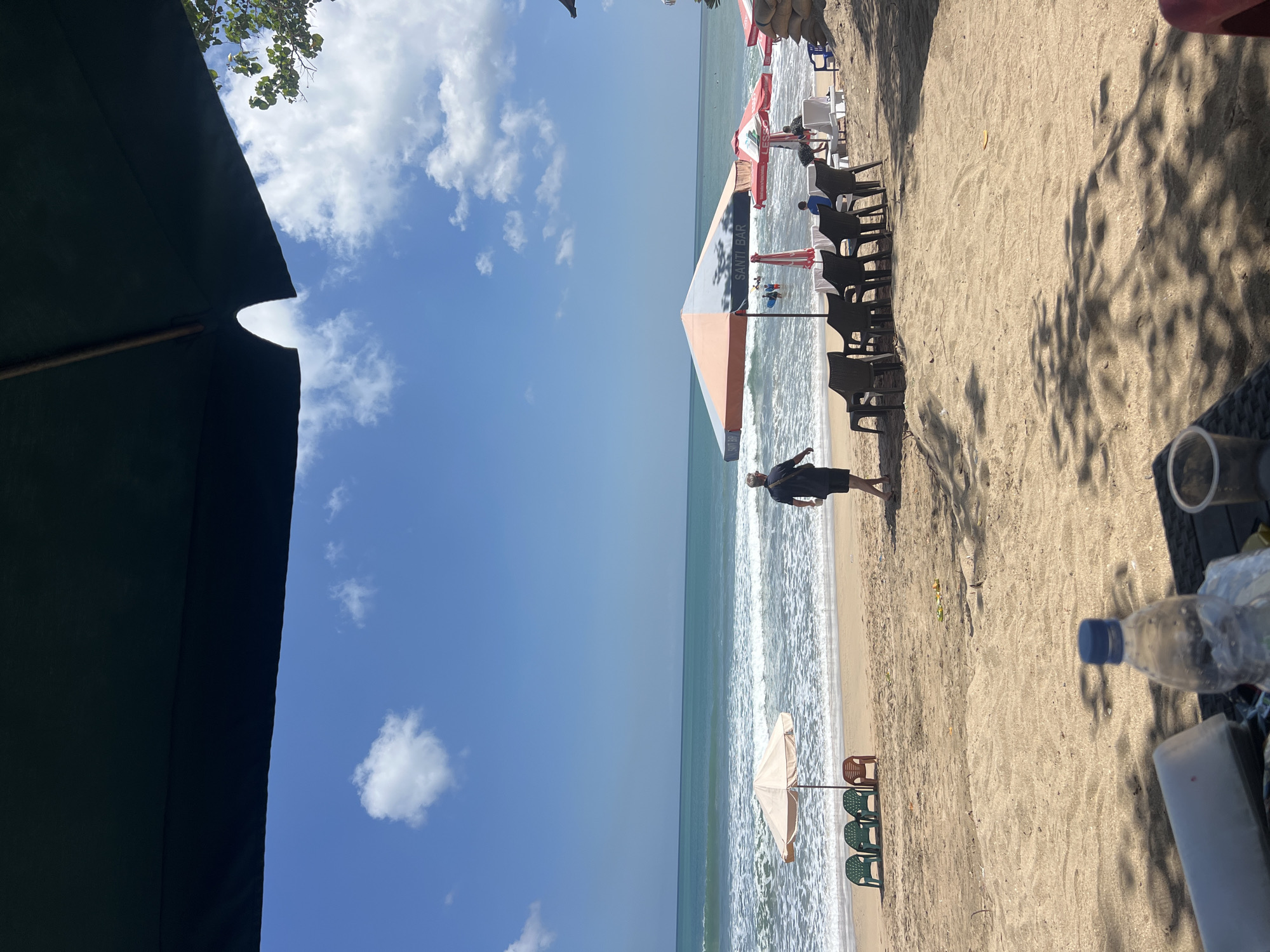 A wide-angle, eye-level shot captures a sunny day at Kuta Beach in Bali, Indonesia, from a shaded vantage point under large umbrellas. The foreground is dominated by sand, with dappled shadows of palm leaves suggesting a tropical setting. A plastic water bottle and other indistinct items rest on a dark surface in the immediate foreground. The middle ground features a sandy beach with rows of empty plastic chairs and a few occupied beach umbrellas, one a large orange and white umbrella with "SANTI BAR" written on it, another a smaller beige umbrella. Several people are visible on the beach and in the ocean, with some wading in the gentle waves and others further out. A man in a dark t-shirt and shorts walks away from the camera towards the ocean. The background is a vibrant turquoise ocean meeting a clear blue sky with scattered white clouds. The overall impression is one of a relaxed, tropical beach atmosphere.