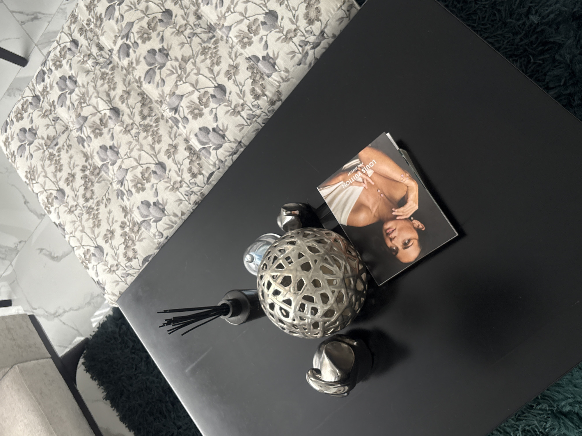 The image shows a black coffee table on a dark green rug. On the table, there is a decorative metal sphere, a diffuser with black sticks, two silver decorative objects, and a Louis Vuitton magazine with a woman on the cover. The upper left corner of the image shows a patterned cushion or ottoman. The location cannot be determined from the image.
