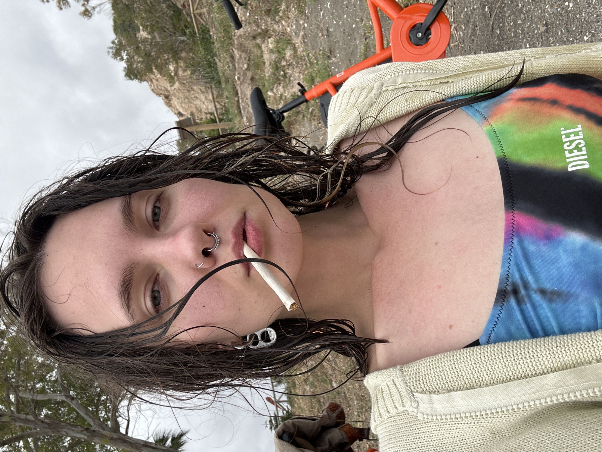 A close-up, rotated portrait shows a young woman outdoors in Málaga, Spain, against a backdrop of an overcast sky and sparse vegetation, with what appears to be a dirt path and an orange and black scooter or bicycle in the background. Her dark, wet-looking hair is prominent, framing her face. She has a septum piercing and a nostril piercing, and an earring resembling a can tab in one ear. A cigarette with a brown filter is held in her mouth, unlit.
She is wearing a colorful top with a tie-dye or abstract pattern in shades of blue, pink, green, and black, featuring the white text "DIESEL" vertically. Over this, she wears a light-colored, possibly cream or beige, knitted cardigan or sweater. Her skin appears slightly flushed. The image is composed as a selfie, with her gaze directed towards the camera. The overall impression is an intimate, casual moment captured outdoors on a cloudy day, possibly near a coastal or natural area given the location context and visible flora.