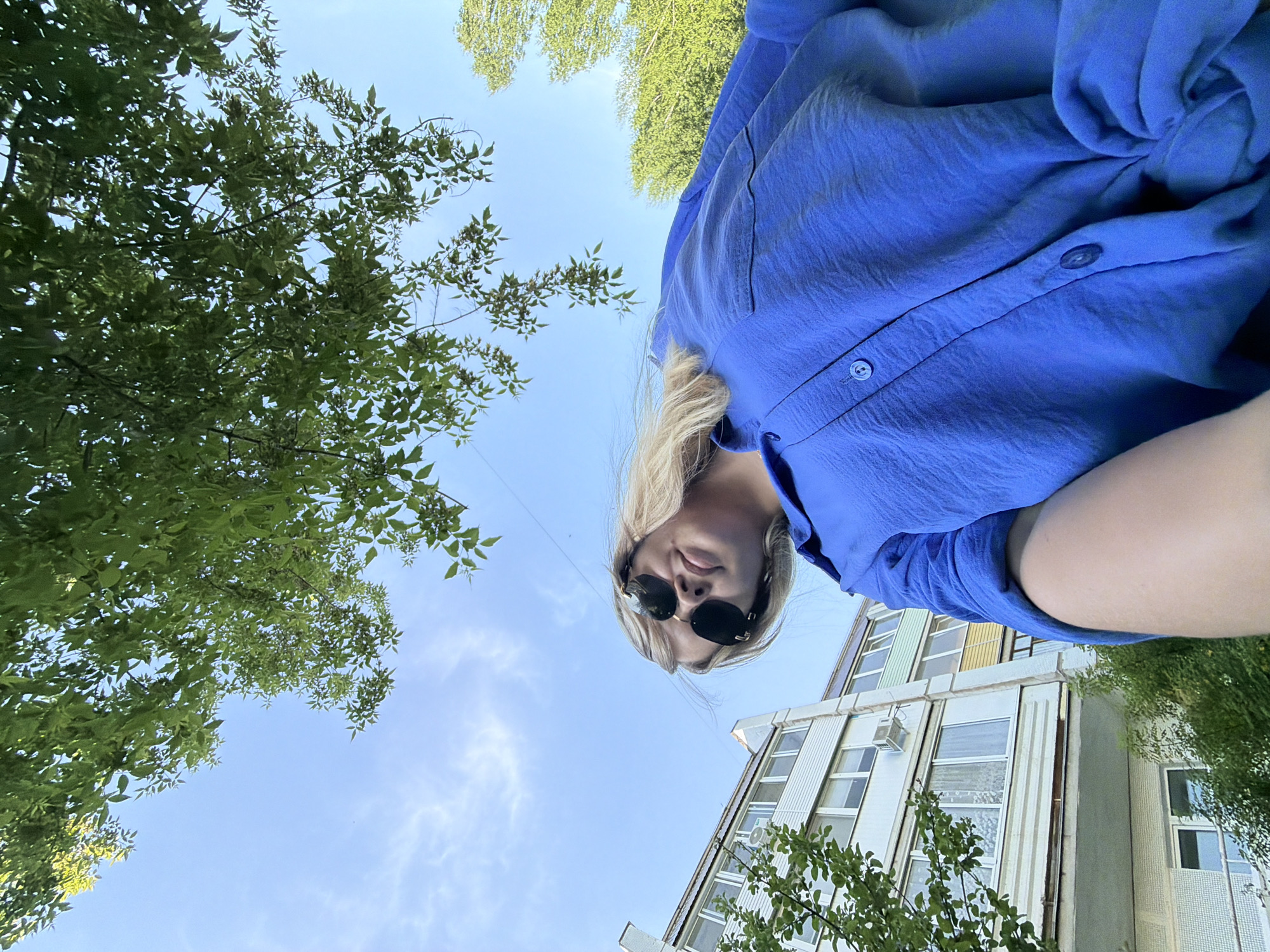 A low-angle shot features a blonde woman wearing sunglasses and a blue collared shirt, positioned above the camera. She is partially obscured by the angle. Behind her is a clear blue sky with scattered clouds, framed by green foliage from trees on either side of the shot. In the lower right corner, a multi-story building is visible with numerous windows and what appear to be air conditioning units attached to some windows.
While identifying the exact location based on a single image is challenging, I cannot pinpoint the precise location in Chaikovsky, Russia, without more contextual information or recognizable landmarks.