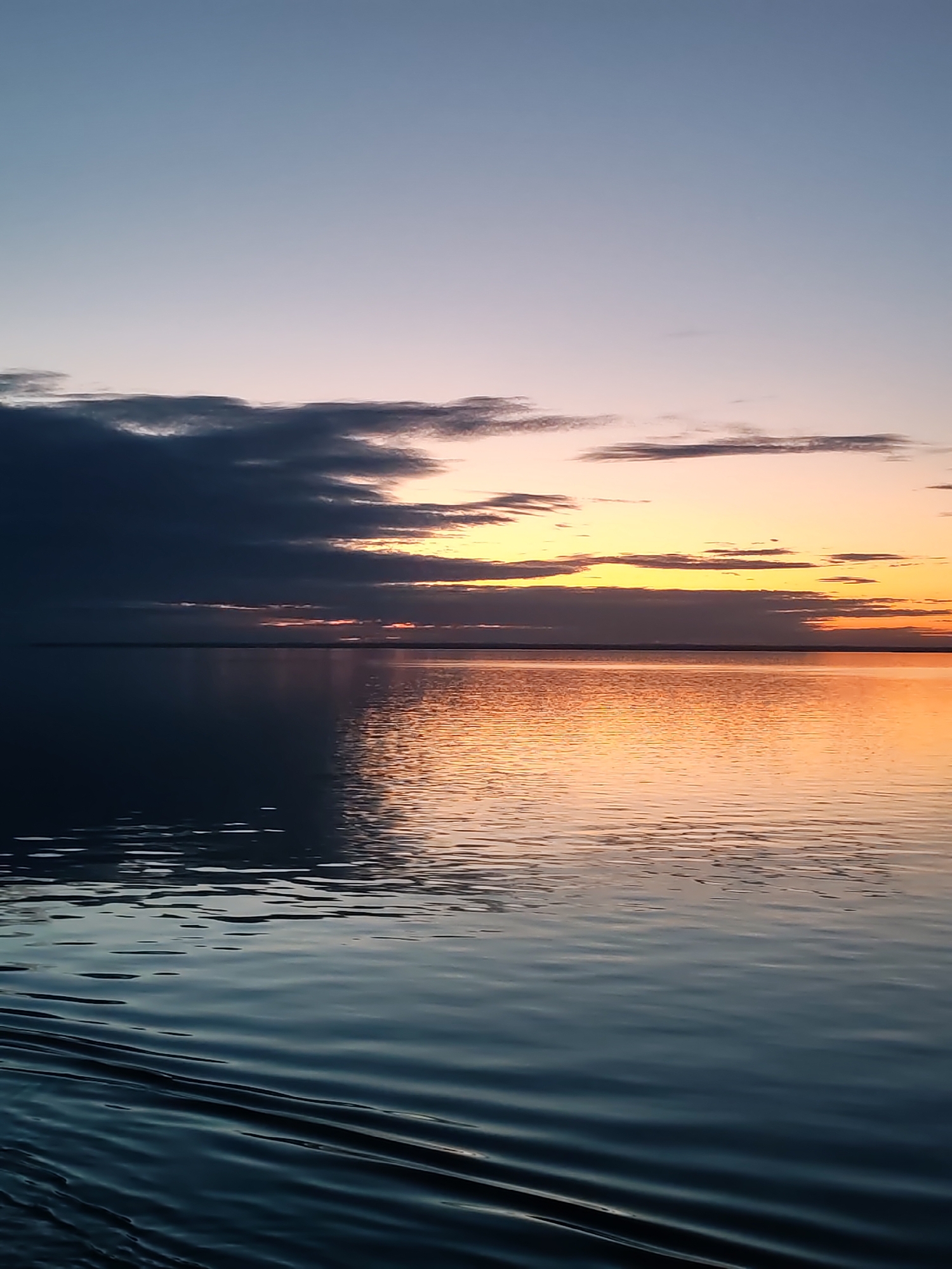 The image presents a serene landscape dominated by a large body of water under a dramatic, colorful sky, indicative of either sunrise or sunset. The sky transitions horizontally, from a soft blue-grey at the top, fading into light purplish-pink and then vibrant orange and yellow hues near the horizon where the sun is low. Dark, heavy clouds are clustered on the left and central parts of the sky, partially obscuring the brightest light but also adding depth and texture. Lighter, wispy clouds are scattered above and within the colorful bands.
The expansive water surface acts as a mirror, reflecting the sky's varied palette. The reflections shift from deep, cool blues and greys in the foreground and on the left to warm, luminous oranges and yellows towards the right, directly under the most intense part of the sky. Gentle ripples and small waves are visible across the water, suggesting a calm environment with a subtle current or breeze. A clear, flat horizon line stretches across the image, separating the vast water from the distant sky, with a faint, low landmass just visible along parts of the horizon.
There are no people, animals, man-made objects, or visible text in the scene, emphasizing the untouched natural beauty. The overall atmosphere conveyed is one of profound tranquility and natural spectacle. The specific location is unknown, but context suggests it is in Russia.