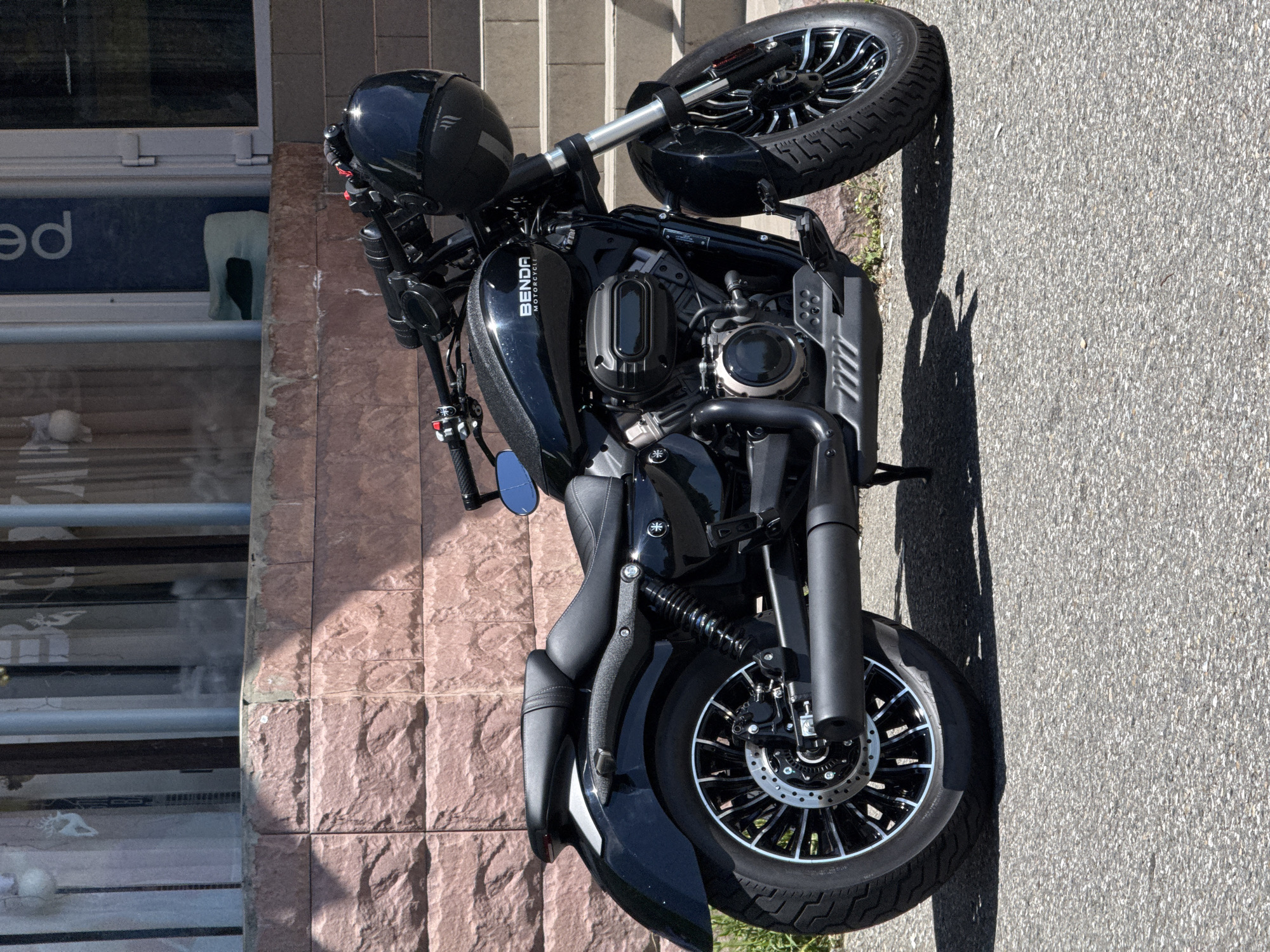 A black motorcycle is parked on an asphalt road next to a building with a textured, reddish-brown stone facade, likely in Sochi, Russia. The image is taken during a bright, sunny daytime, casting distinct shadows on the ground from the motorcycle and nearby objects.
The motorcycle is a cruiser-style bike, predominantly black with some silver accents on the engine components and wheel spokes. The brand name "BENDA MOTORCYCLE" is visible in white lettering on the glossy black fuel tank. The engine appears to be a V-twin with prominent black covers. The front wheel features intricate black and silver multi-spoke rims with a disc brake. A black motorcycle helmet rests on the handlebars.
To the left of the motorcycle, a window is visible in the building, reflecting parts of the surroundings. Inside the window, a sign or poster is partially visible with the letters "ed" in white on a blue background. The overall scene suggests the motorcycle is temporarily parked, perhaps by a building entrance or storefront.