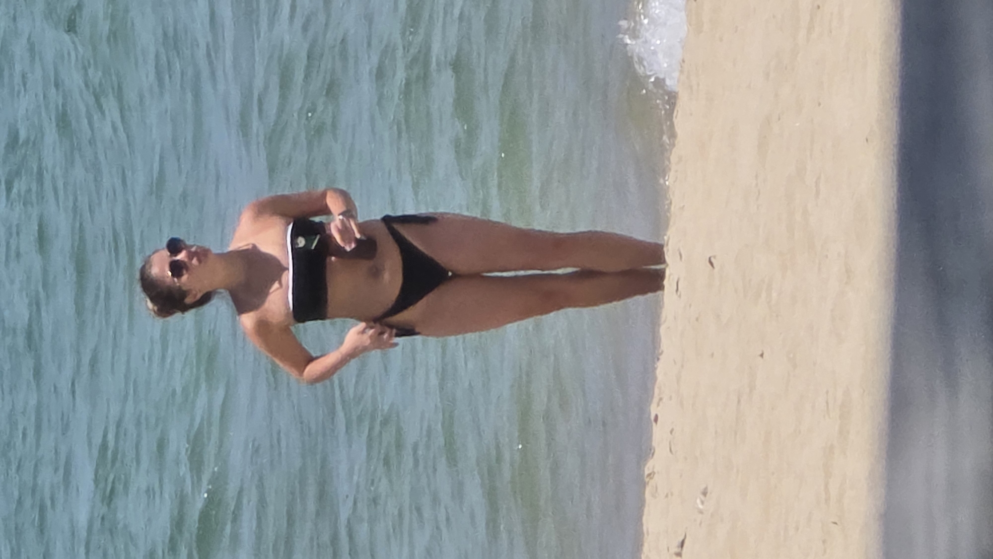 A woman in a black bikini stands on the beach at Nha Trang, Vietnam. She holds a black object, possibly a phone, in her left hand and wears sunglasses. The beach sand is light beige, and the ocean water is a light turquoise. Waves are visible near the shoreline.