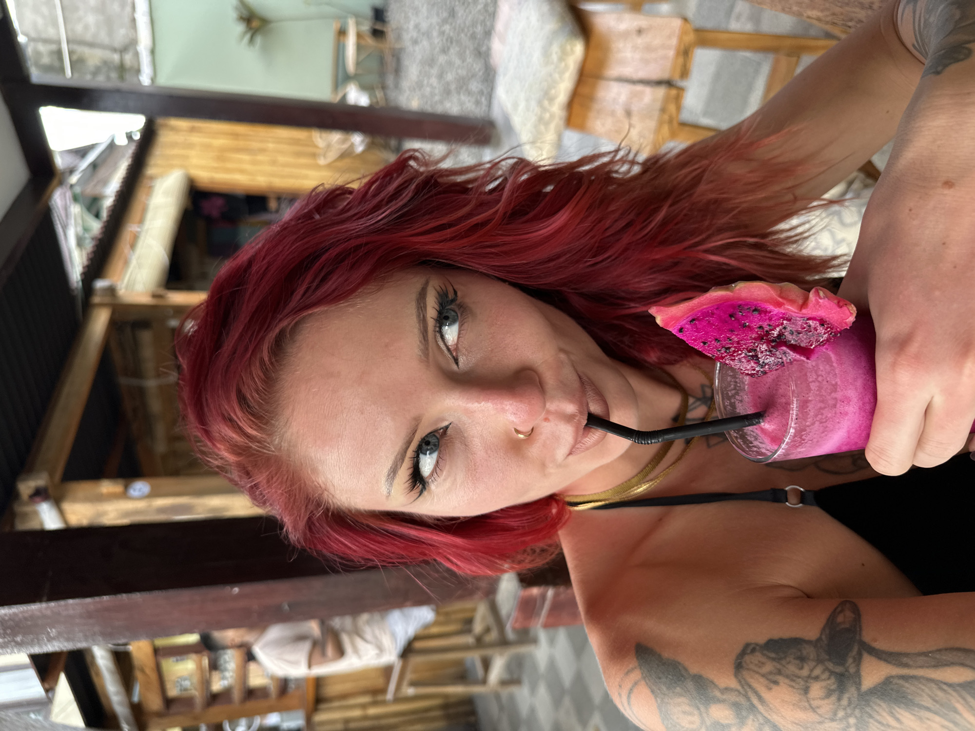 A woman with red hair is shown from the chest up, drinking a pink smoothie with a black straw. A slice of dragon fruit is perched on the glass. She has a gold nose ring and a gold necklace. Tattoos are visible on her right arm. The background shows a partially visible outdoor seating area. The location is Unknown, Indonesia.