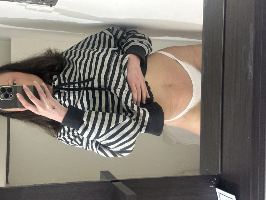The scene depicts a female individual taking a mirror selfie indoors, in a setting consistent with a bathroom, located in Tunbridge Wells, United Kingdom. The individual is shown from approximately the upper chest to mid-thigh, reflected in a mirror. She has dark hair and wears a dark and light grey horizontal striped hooded sweatshirt, unzipped to expose her midriff. White thong-style underwear is visible. Her right hand, holding a dark-colored smartphone, obscures the majority of her face. A small, dark, star-shaped tattoo is visible on the back of her right hand, near the index finger knuckle. Her left hand rests on her midriff, which displays visible skin marks consistent with stretch marks. The background, reflected in the mirror, includes light-colored walls, a partially visible white toilet bowl behind the individual's lower torso, a dark vertical shelf-like structure on the left, and a dark rectangular fixture mounted on the wall to the right. The bottom foreground shows the top edge of a dark brown mirror frame or vanity surface.