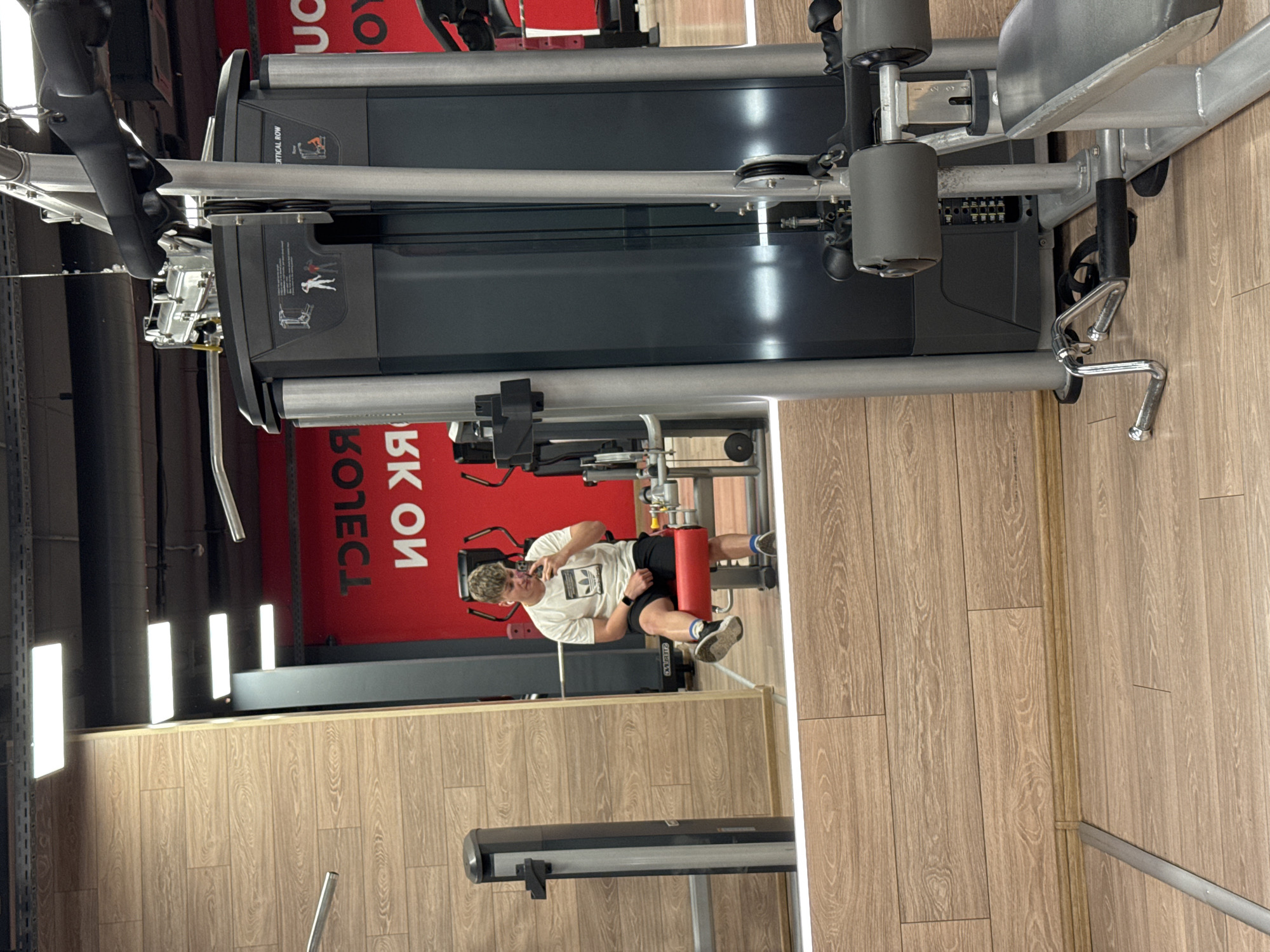 The image displays the interior of a gym, likely a Pulse Fitness & Spa facility in Plovdiv, Bulgaria, indicated by the prominent red wall with reversed white text in the mirror reflection, characteristic of the brand's aesthetics (e.g., "WORK NOW" or "PULSE" in reverse).
The primary subject is a young adult male, with short, light-colored hair, captured in a mirror reflection. He is wearing a white t-shirt with a black Adidas logo, black athletic shorts, white socks with blue accents, and dark athletic shoes. Wireless earbuds are visible in both ears, and an Apple Watch is on his left wrist. The male is seated on the red padded leg roller of a leg extension machine, holding a dark-cased smartphone in his right hand, angled to take a mirror selfie. His left hand rests on his left thigh.
To the right of the frame, a grey and black cable machine or multi-gym station is partially visible, featuring a seat, padded rollers, and various cable attachments. A curved chrome handle accessory rests on the light brown wood laminate floor near the base of this machine. The floor throughout the visible area is light brown wood laminate. The wall on the left side of the frame is wood-paneled. The ceiling features exposed dark structures and multiple rectangular white LED light fixtures.