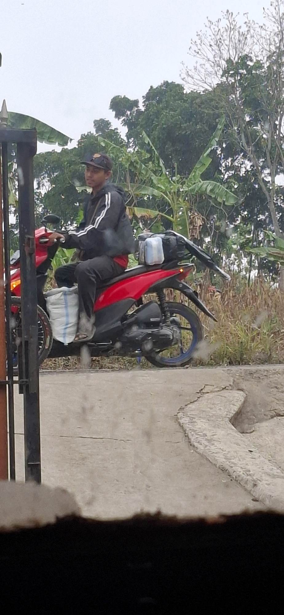 A person wearing a black jacket with white stripes, dark pants, and a baseball cap rides a red motorcycle. The motorcycle has a white bag secured to the front and a black bag with two blue containers on the back. The rider is looking to their left, and the motorcycle's exhaust is emitting smoke. The scene appears to be in a rural or semi-rural area with trees and vegetation in the background, and a cracked concrete path in the foreground. The sky is overcast. The image is likely taken in Cianjur, Indonesia, given the context.