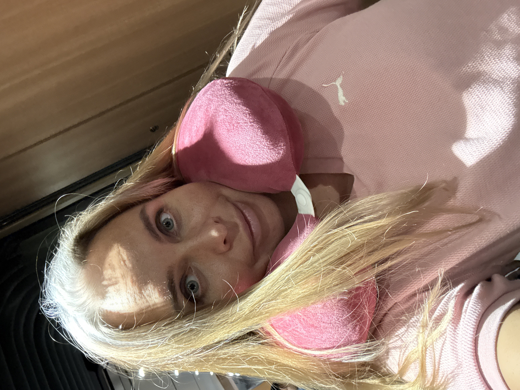 A woman is posing for a selfie, looking towards the camera with a smile. She has blonde hair that is partially grey at the roots. She is wearing a pink sweatshirt with a small white Puma logo on the left chest. Around her neck is a pink neck pillow. The background appears to be an indoor setting, possibly a vehicle or a waiting area, with wooden paneling on the right side. The image is lit by overhead lighting, casting shadows on the woman's face and clothing. The woman is positioned in the foreground, with her head tilted slightly to the left.