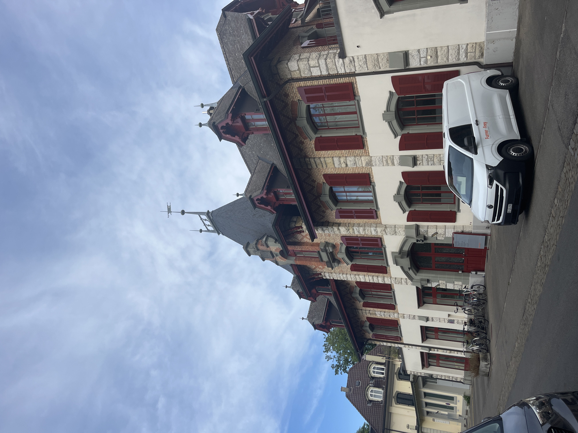 This image shows the exterior of a multi-story building in Bern, Switzerland. The building has a classical architectural style with red shutters on the windows and a dark, steeply pitched roof with dormer windows. A white van with "Furrer + Frey" written on its side is parked in front of the building. Several bicycles are also parked in front of the building. The sky is partly cloudy. The image appears to have been taken from street level, with a car visible in the foreground.