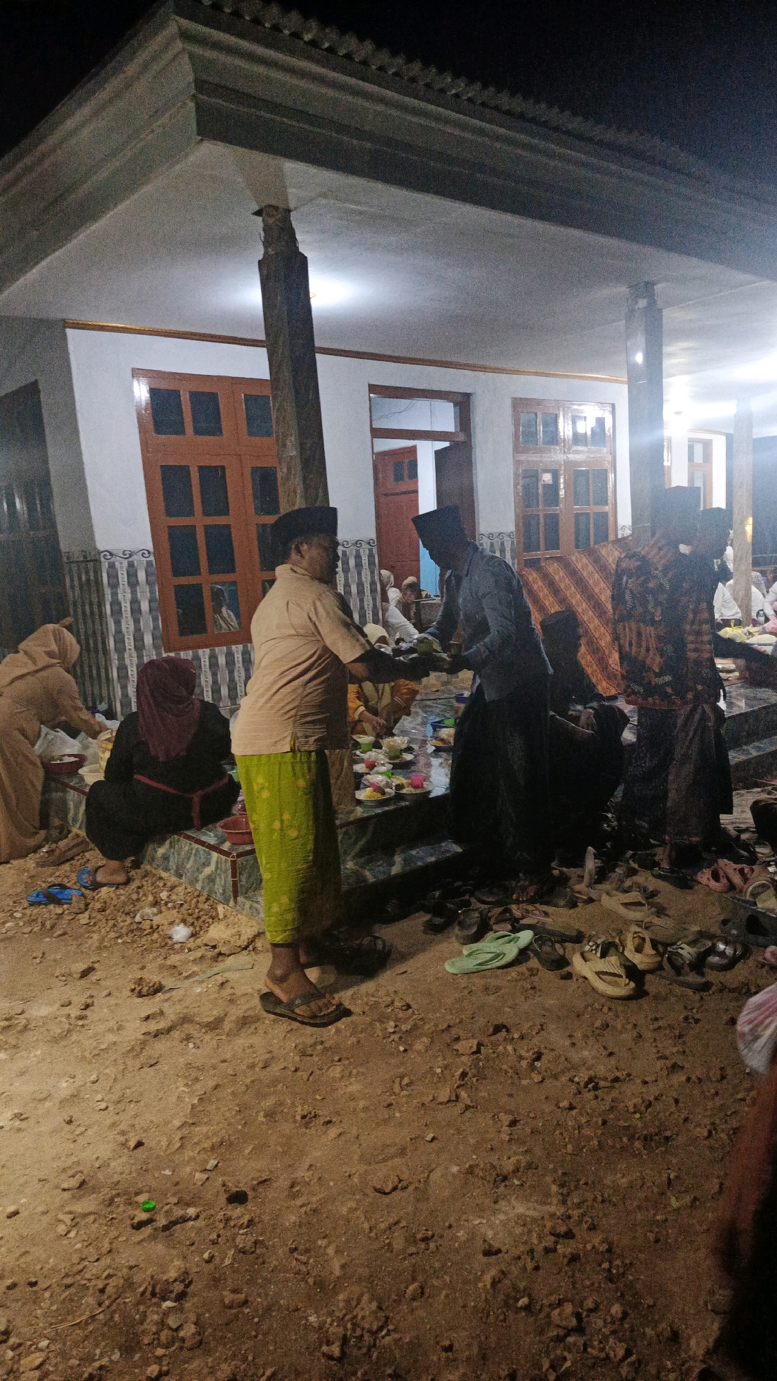 In Sokaramme Timur, Indonesia, a group of men and women are gathered outdoors at night, possibly for a religious or cultural event. The scene is dimly lit, with artificial lights illuminating the area. Several individuals are dressed in traditional Islamic attire, including peci hats and sarongs. Some men are seen in the process of serving or receiving food from plates arranged on a raised platform. Others are seated or standing, engaged in conversation or observation. A few women, also in traditional dress, are visible in the background. The ground is unpaved and appears to be dirt or sand, with scattered rocks and debris. Numerous pairs of sandals and shoes are discarded on the ground, indicating that participants have removed them before entering the gathering area. The architectural elements suggest a building with a tiled roof and wooden window frames.