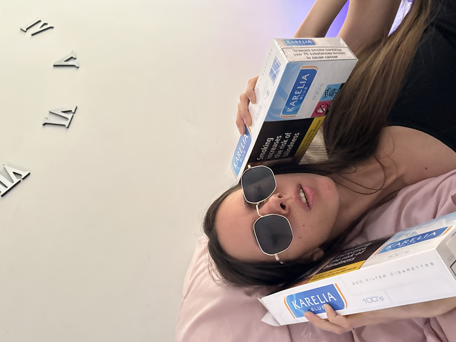 The scene depicts a female individual, appearing to be an adult, reclining on a light pink pillow or bedding in an indoor setting. She has long, dark brown hair and is wearing polygonal-shaped sunglasses with dark lenses and a gold-colored frame, along with a dark-colored top. Her mouth is slightly ajar. She is holding two large cartons of "KARELIA BLUE 100's" filter cigarettes, one on each side of her head. The carton on the viewer's right prominently displays the UK health warning "Smoking increases the risk of blindness" and a graphical warning of a pregnant woman with a crossed-out symbol. The carton on the viewer's left is partially visible, also showing "KARELIA BLUE 100's filter cigarettes." The background consists of a plain white wall, on which several black Roman numeral clock markers are affixed, specifically "XII," "II," and "IV." A subtle purple ambient light emanates from the right side of the wall. The specific location within Corby, United Kingdom, cannot be determined from the visual evidence provided; however, the cigarette packaging confirms a UK context.