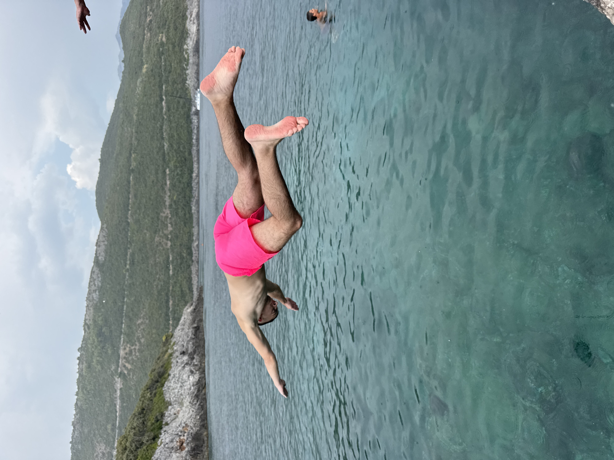 A person is captured mid-air, diving into the clear turquoise waters of the Adriatic Sea off the coast of Dubraljevina, Montenegro. The individual, wearing bright pink swim shorts, is positioned horizontally with arms extended forward and legs bent backwards. The rocky coastline with sparse green vegetation forms the backdrop, leading to a mountainous terrain under a cloudy sky. A white sailboat is visible in the distance, and another person is swimming in the sea. The water is calm with gentle ripples, and the sunlight casts a slight reflection on its surface.