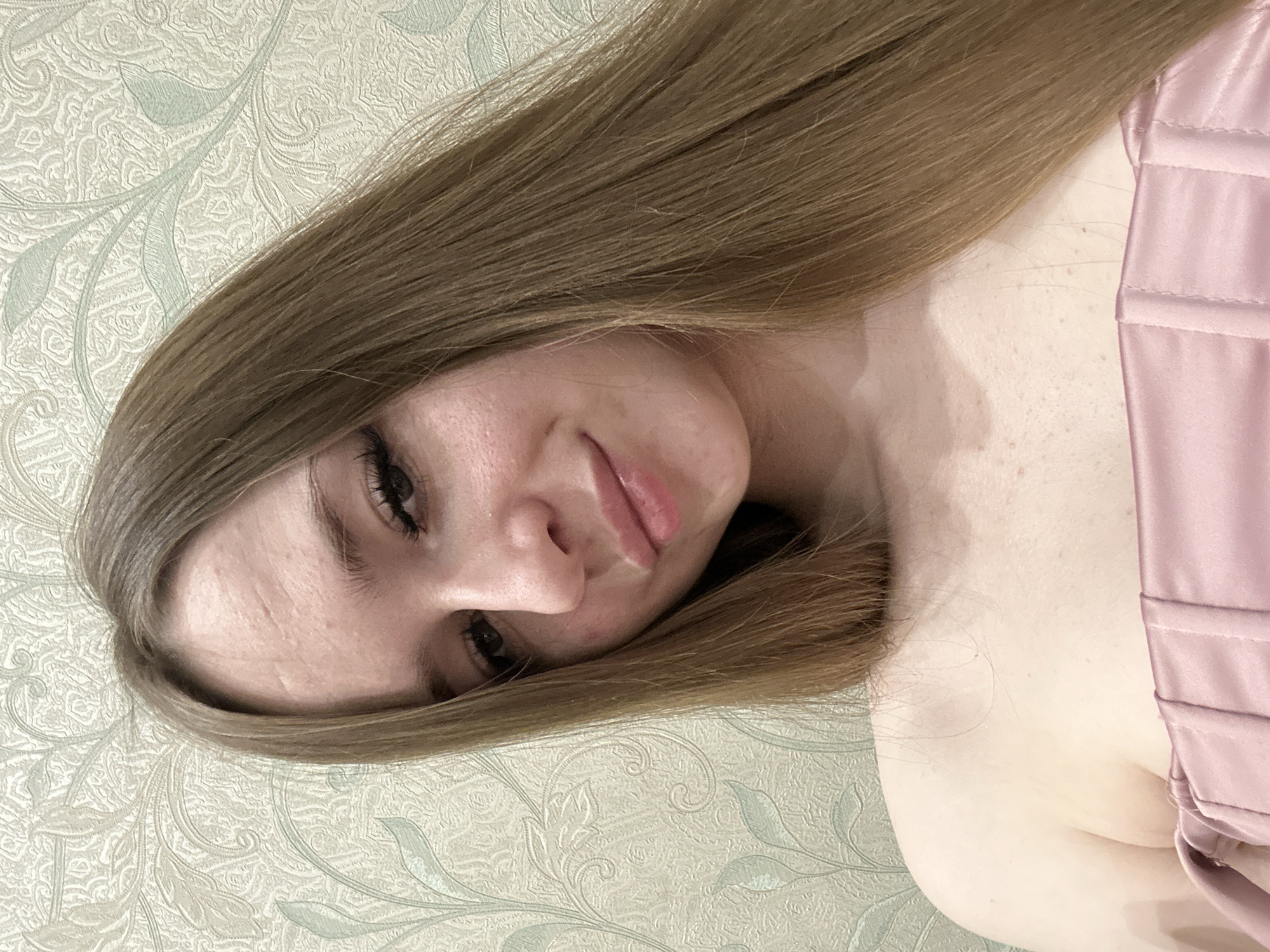 A close-up, rotated portrait photograph shows a young woman with long, straight blonde hair. She is fair-skinned and wears dark mascara. She has a slight smile. She is wearing a pale pink corset-style top. The background is a patterned wallpaper with a light green and beige color scheme. The location and city are not identifiable from the image.
