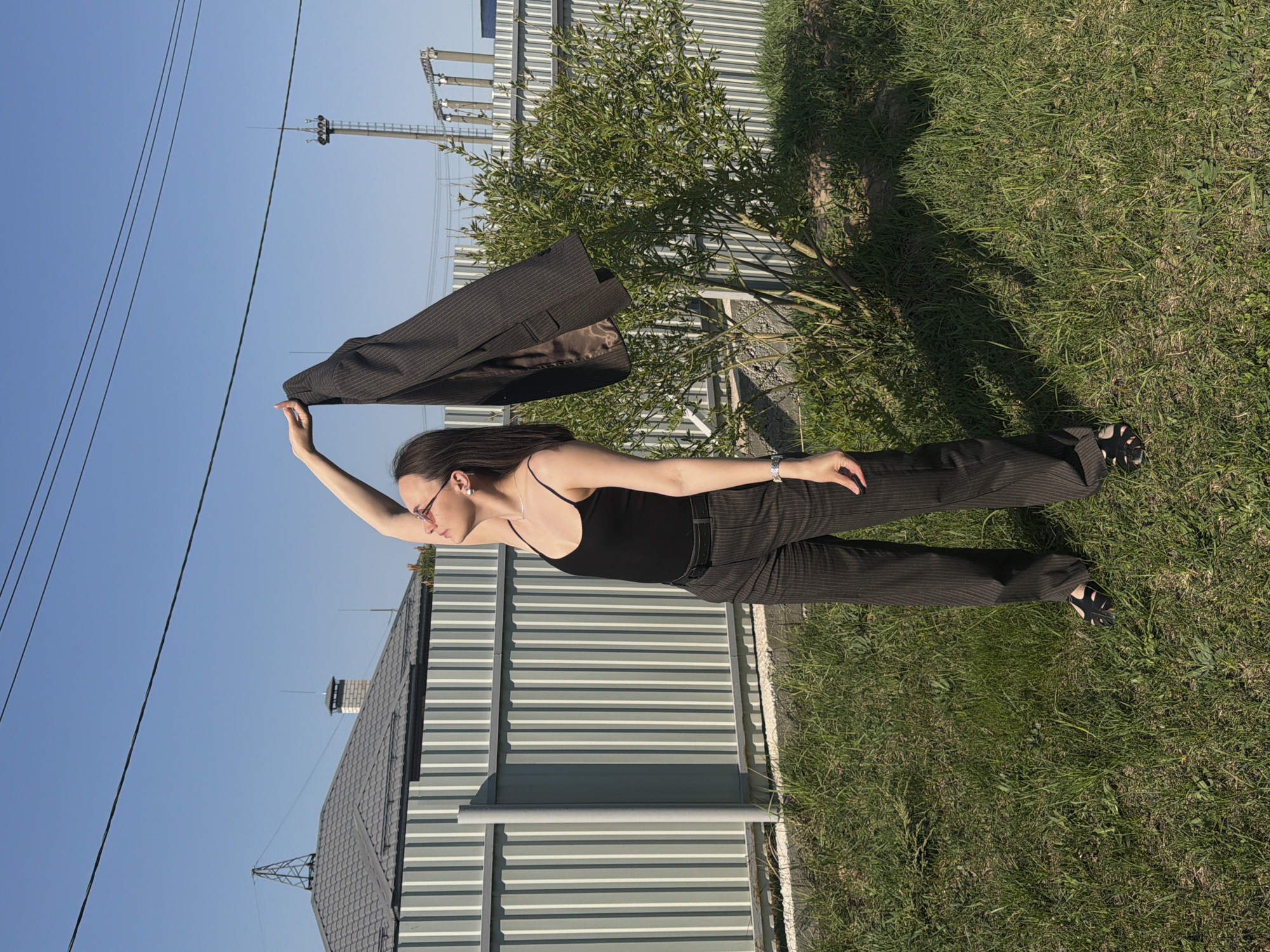 The image shows a woman standing outside, on grass, with a gray, corrugated-metal fence and building behind her. She is wearing a black tank top, dark pinstripe pants, sandals, and glasses. She is holding a dark pinstripe blazer over her head, with her right arm extended straight up. In the background are some utility poles, transmission towers, trees, and another building with a gray roof. The sky is clear blue. The location is Chaikovsky, Russia.