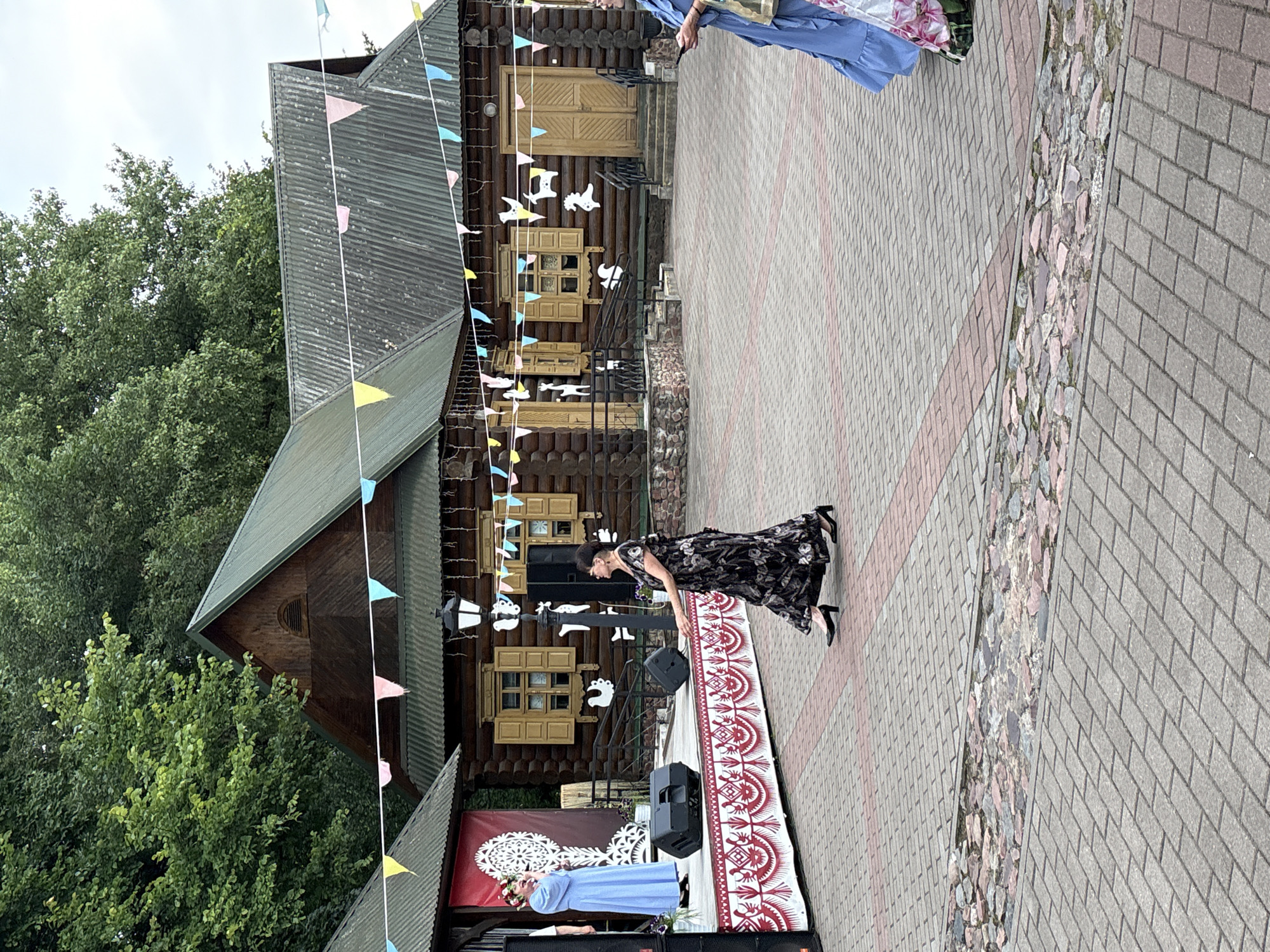 The image depicts an outdoor performance space. In the background is a rustic wooden building with a corrugated metal roof, decorated with colorful triangular flags. In the foreground, a woman in a black floral dress is captured mid-dance on a paved courtyard. To her left, a woman in a blue dress stands on a stage, singing into a microphone. The stage is adorned with red and white decorative trim and flanked by speakers. The overall setting suggests a cultural event or festival. The specific location of this scene is likely within the Vitebsk City Open-Air Museum of Traditional Architecture, located in the city of Vitebsk, Belarus.