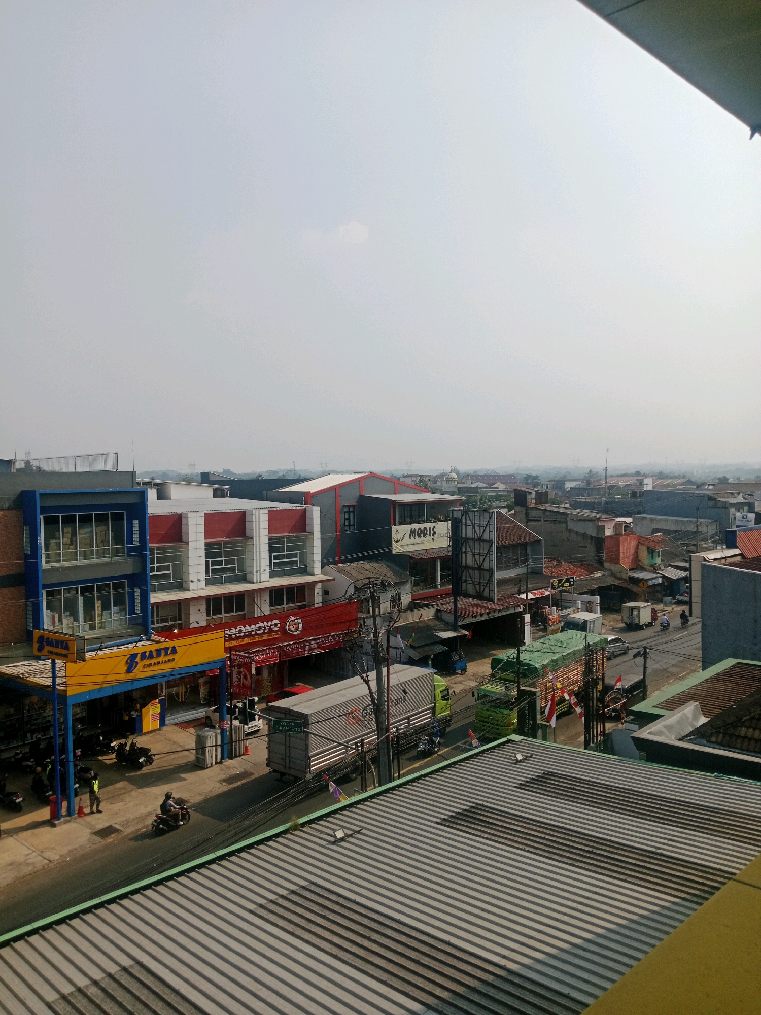 The image is a daylight, medium-angle, outdoor shot of a street scene in Ciranjang, Indonesia. The foreground is dominated by the corrugated metal roof of a building. Beyond the roof, a two-lane street runs horizontally across the frame, bordered by buildings on both sides. On the left side of the street, there are multiple commercial establishments. One building, painted blue and gray, has large glass windows displaying products. Adjacent to it is a yellow awning with the text "SANTYA CIRANJANG". Further down the street, a red awning displays "MOMOYO" with smaller text "ICE CREAM FRUIT TEA SUNNY". Several motorcycles are parked on the sidewalk in front of these shops. A few people are visible, including someone in a black jacket standing near a red cone on the sidewalk, and a person riding a motorcycle.
On the right side of the street, various buildings of different architectural styles and colors are present. Some appear to be residential, while others are commercial. A white truck with a large cargo container, marked with "YOGYA CIRANJANG", is stopped on the street. Several other vehicles, including trucks and cars, are also present. A few Indonesian flags, red and white, are displayed on poles attached to some of the vehicles. The background shows a hazy, urban landscape with more buildings and some vegetation, extending towards the horizon. The sky is overcast with a light haze.