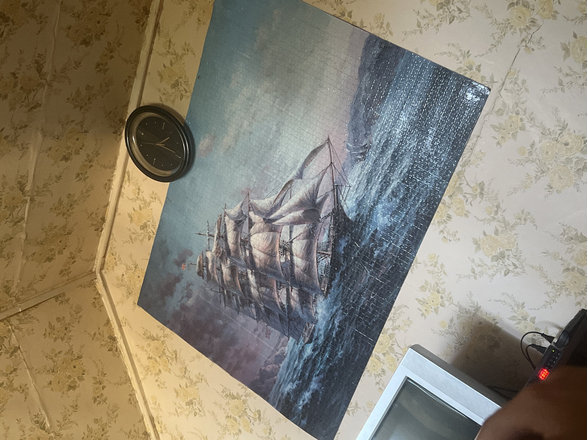 The image shows a partially completed jigsaw puzzle depicting a tall ship sailing on a rough sea under a dramatic sky. The puzzle is laid out on a surface covered with floral patterned wallpaper. A round, black analog clock with white hands is placed on the puzzle, near the top right corner. In the bottom left of the image, a white electronic device with cables and a small red digital display is visible, along with a hand holding a dark object. The scene appears to be indoors, with the floral wallpaper extending onto the ceiling, suggesting a room. There is no visual information in the image that can be used to determine the location in the city of Krasnoslobodsk, Russia.