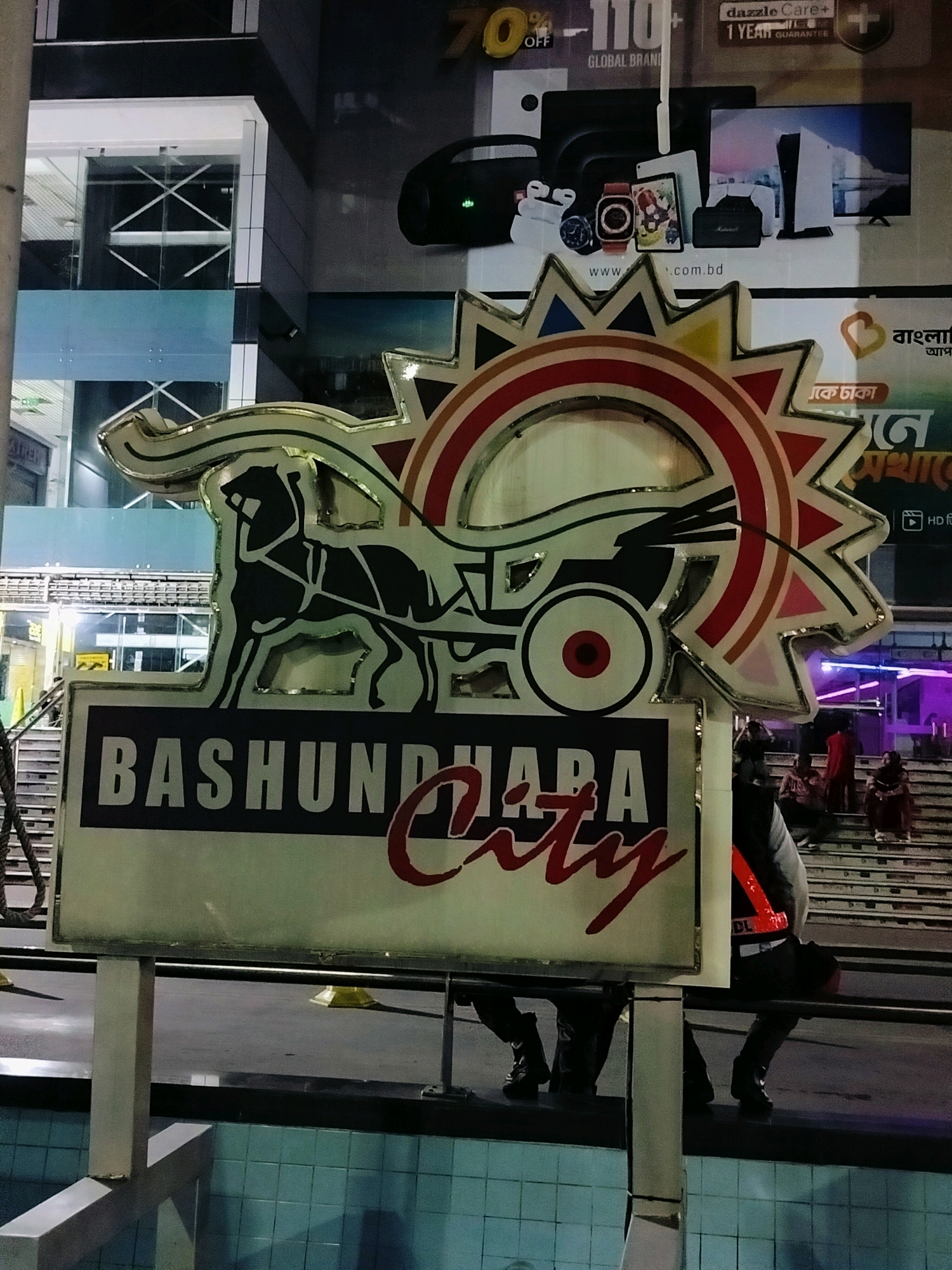 The image shows a sign for "BASHUNDHARA CITY" in Dhaka, Bangladesh. The sign features a stylized sun with a horse-drawn carriage. In the background, there are people walking up stairs. The sign is illuminated and appears to be in a public area. The building behind the sign is modern, with glass windows. The overall scene is a nighttime street view in a commercial area.