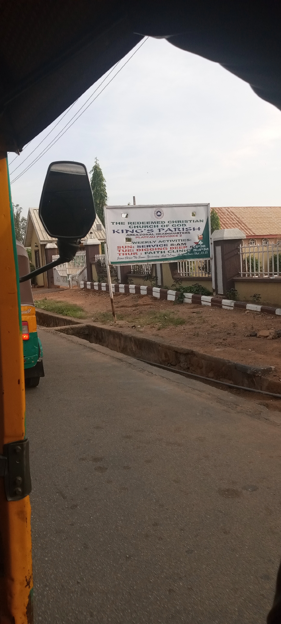 The image shows a street view of The Redeemed Christian Church of God, King's Parish, Area/Zonal Headquarters in Jos, Nigeria, taken from inside a vehicle, possibly a taxi or "keke napep". A sign in front of the church lists weekly activities: Sunday service at 8 AM, Digging Deep on Tuesday at 5 PM, and Faith Clinic on Thursday at 5 PM. The environment includes the street, a curb, the church building with a fence, and some landscaping.