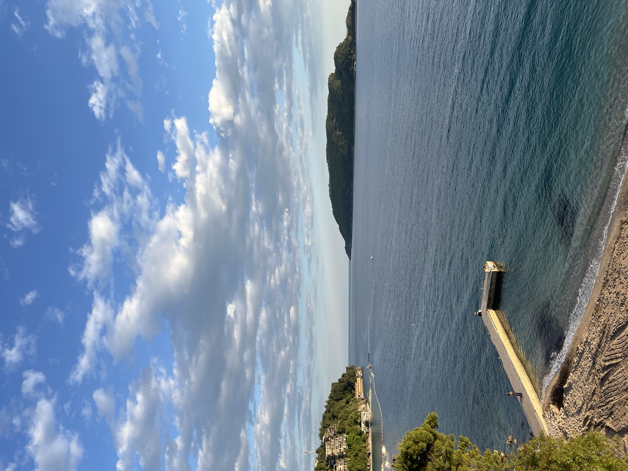 The image presents a coastal scene in Budva, Montenegro, viewed from an elevated perspective. The sky is dominated by puffy white clouds against a blue backdrop. Below, the Adriatic Sea is a deep blue, stretching towards a densely vegetated island in the distance. Closer to the foreground, a concrete pier extends into the water, with one individual standing on the end. The shoreline is sandy, and waves are lapping on the beach. Adjacent to the beach is a landscape with greenery.