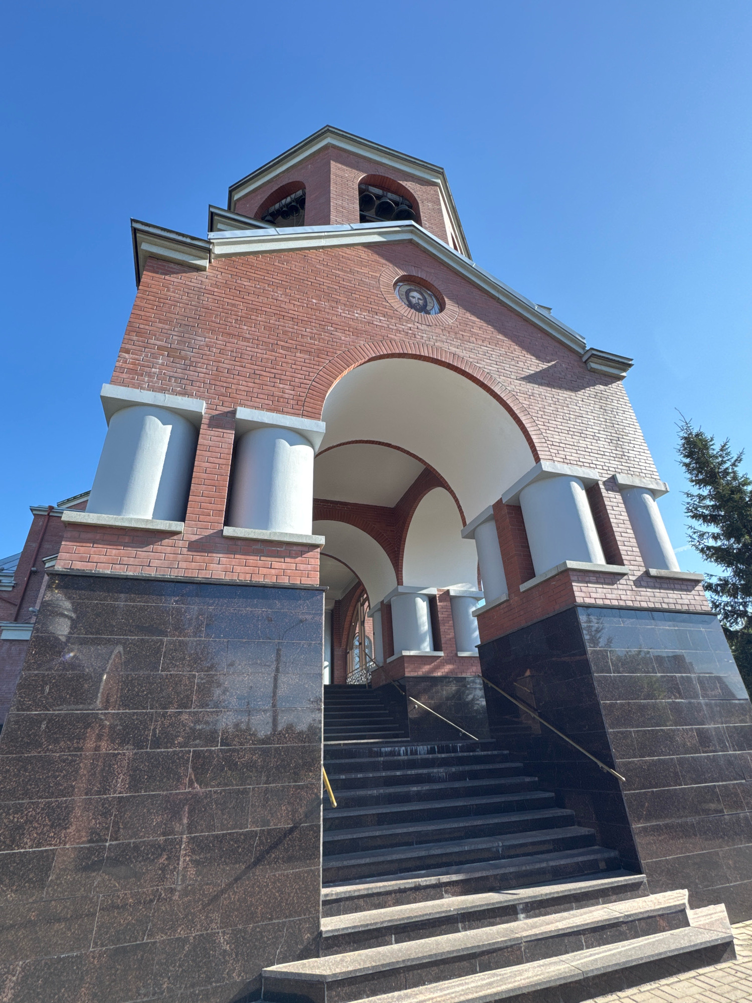 The image shows the entrance of a church in Saint Petersburg, Russia, with a clear blue sky in the background. The church is constructed of red brick and features arched entrances and white columns. The entrance is accessed by a wide set of stairs. The base of the church and the stairs are clad in dark granite. A tall evergreen tree is visible on the right side of the image. The perspective is low-angle, looking up at the structure.