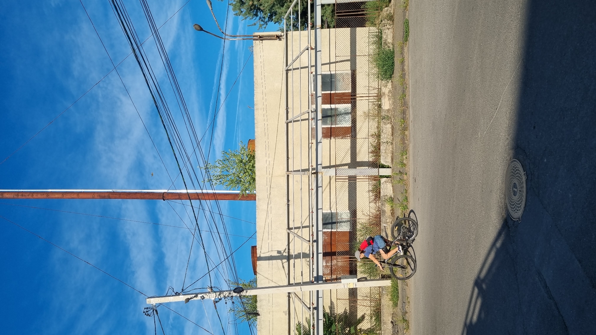 A person rides a bicycle past an industrial-looking building with a tall, rusty pole and numerous overhead wires against a bright blue sky with wispy clouds. The building is light beige and appears to have several windows covered with brown material. A chain-link fence runs along the front of the building. The bicycle rider is wearing a blue shirt, dark shorts, a grey cap, and a red backpack. The bicycle is a dark color, possibly black or dark grey. The road in the foreground is asphalt and a manhole cover is visible. The overall scene suggests an urban or semi-industrial setting in Berehove, Ukraine.
