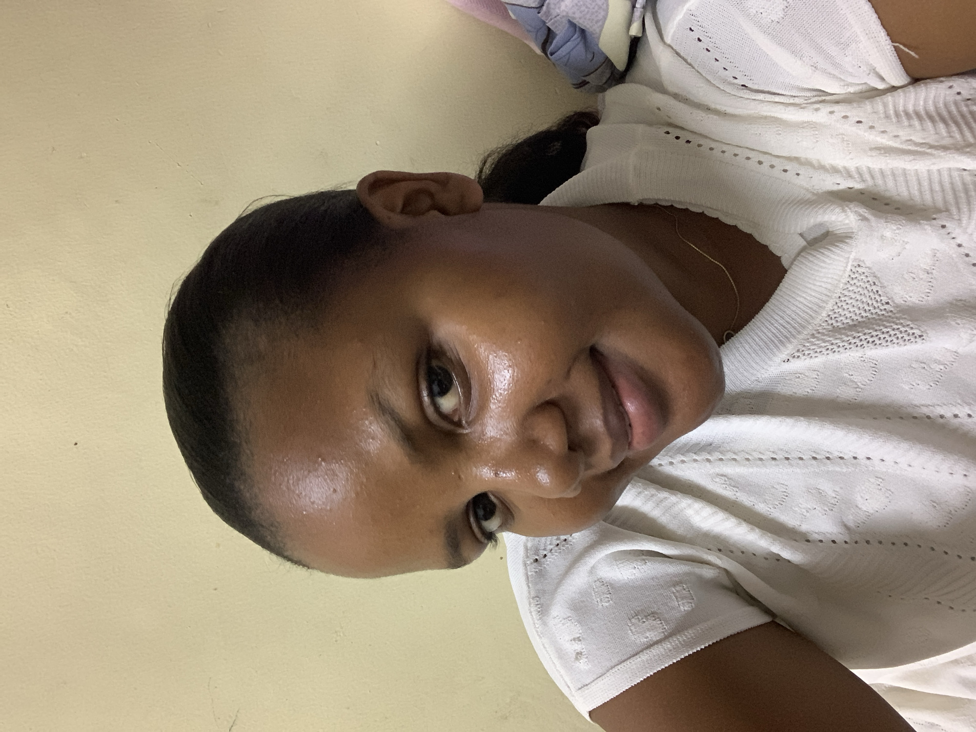 A close-up, rotated image depicts a person with dark hair pulled back, brown skin, and dark eyes. They are wearing a white patterned sweater. The background is a pale yellow wall. There is no indication of the location being in Dar es-Salaam, Tanzania.