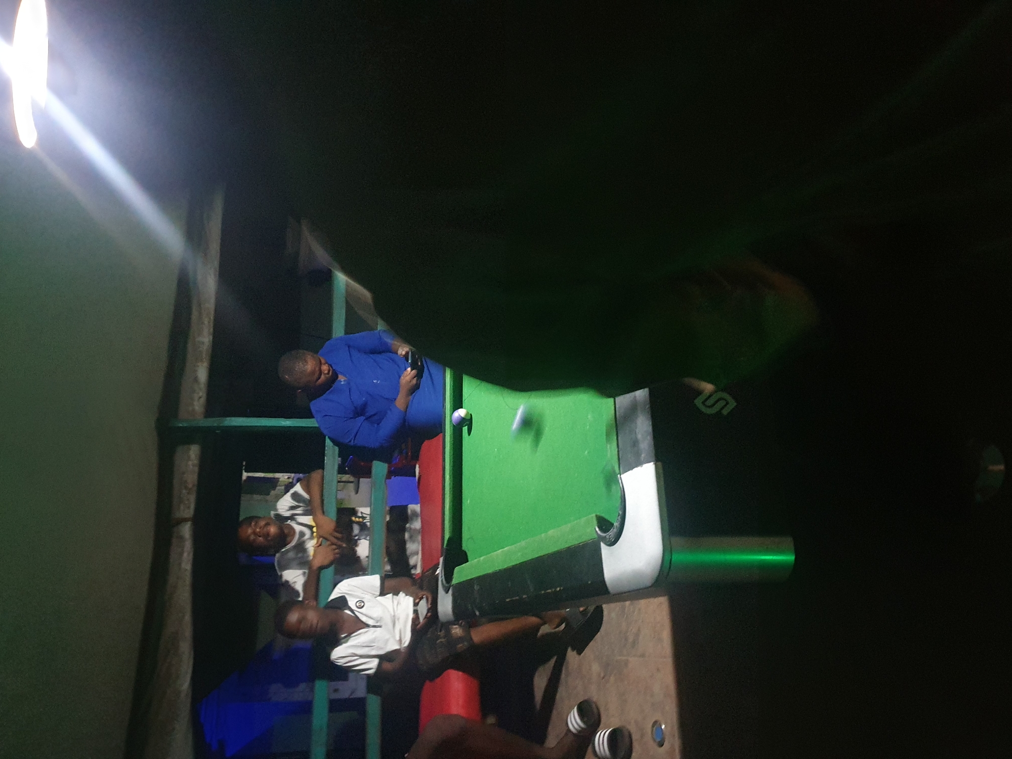 In Lokoja, Nigeria, a group of young men are gathered around a green billiard table. Two of the men are seated, one in a white polo shirt and patterned shorts, and the other in a white and grey patterned t-shirt. Another man, wearing a blue shirt, stands near the table, holding a mobile phone. The billiard table has brightly lit green legs, and a cue ball is in motion on the table's surface. The scene appears to be outdoors, possibly at a bar or recreational area, with a roof overhead and a bright light fixture illuminating the space. The lighting suggests it is evening or nighttime.