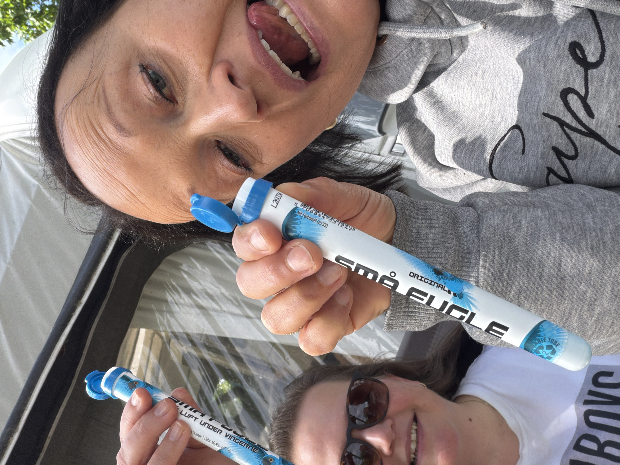 A selfie-style photograph depicts two adult women outdoors in what is identified as Horten, Norway.
The foreground subject, an adult woman with dark hair, occupies the right half of the frame. She wears a grey hooded sweatshirt with black embroidered text, partially visible as "supe". Her mouth is wide open with her tongue slightly extended in a playful or exaggerated expression, and her eyes are directed towards the camera. In her right hand, she holds a white, cylindrical "Små FUGLÆ ORIGINAL" liquor tube (16.4% Vol.) with an open blue cap, featuring a blue bird motif. The tube's label also displays a batch number "L28004" and a barcode.
Behind her, partially obscured on the left, an adult woman with lighter hair and dark sunglasses smiles towards the camera. She wears a white t-shirt with dark text visible as "BOYS" at the bottom left. She holds a similar "Små FUGLÆ" tube in her left hand, which is less clearly visible.
The background features a white tent or awning structure with a clear plastic window panel that reflects light and shows distant green foliage and blue sky. A patch of clear blue sky is also visible at the upper right. The scene is brightly lit by natural sunlight, indicating daytime.