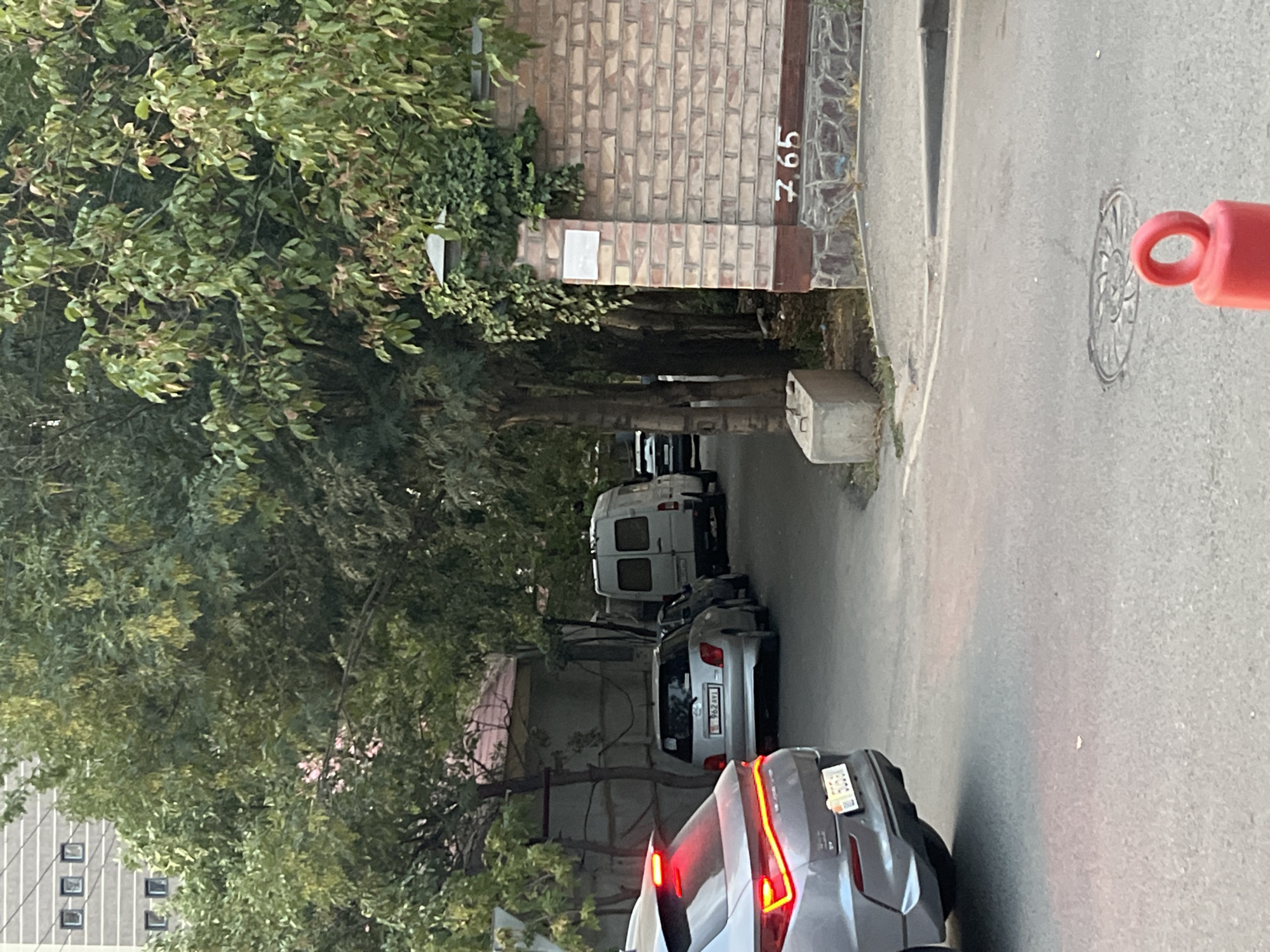 The image shows a street scene in Bishkek, Kyrgyzstan. In the foreground, the rear of a silver SUV is visible, with its brake lights illuminated. A narrow paved road curves to the right, with a concrete curb. Further down the road, several parked vehicles are visible, including a silver hatchback and a white van. Trees line the street, and a brick wall with the number "765" painted on it is present on the right side of the image. A concrete block is on the side of the road near the brick wall. A manhole cover is visible in the road. The overall impression is that of a residential or semi-industrial area.