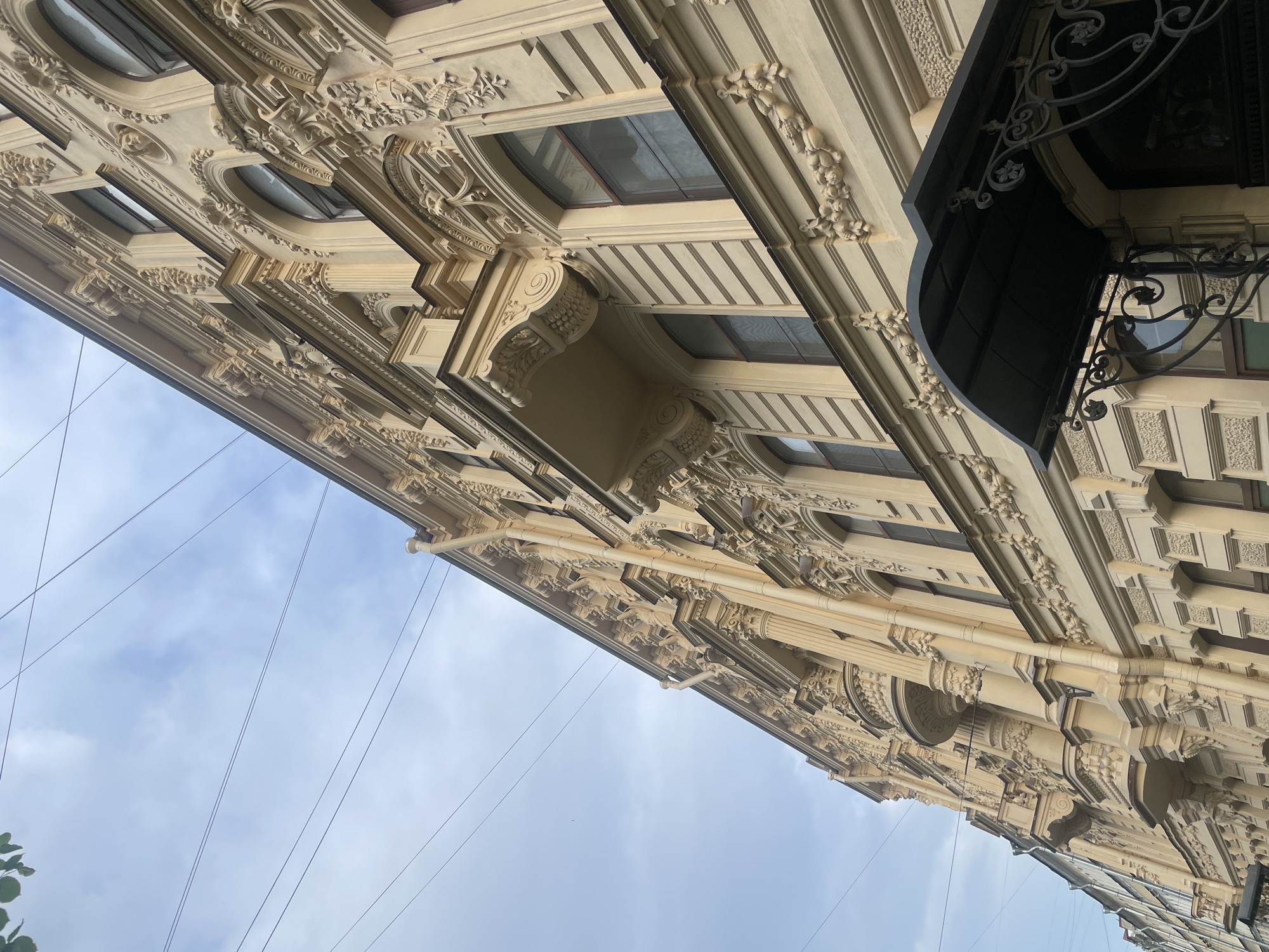 The image provides an upward-angled view of a highly ornate building facade in Saint Petersburg, Russia, set against a partly cloudy sky. The building, likely made of light-colored stone or stucco, exhibits a rich and intricate architectural style, possibly Neoclassical or Art Nouveau, characterized by abundant decorative elements.
The facade is adorned with multiple levels of elaborate detailing, including richly sculpted cornices, decorative moldings, pilasters, and volute-shaped corbels. Oval and rectangular windows are integrated into the design, many framed by intricate surrounds. Reliefs depicting foliage, garlands, and possibly stylized figures are visible along friezes and other architectural components, showcasing fine craftsmanship.
A prominent feature is a dark metal awning or canopy with delicate wrought-iron scrollwork, projecting from the upper right side of the building. A vertical downspout or pipe runs down a section of the facade. Thin black power lines crisscross the upper left portion of the frame against the sky, indicating an urban environment. A hint of green foliage is visible in the bottom left corner.
The time of day appears to be daytime, with diffuse light from the overcast or partly cloudy sky illuminating the building's details and creating subtle shadows that enhance its three-dimensional quality. There is no visible activity or explicit text. The image emphasizes the grandeur and detailed artistry of the historic architecture.