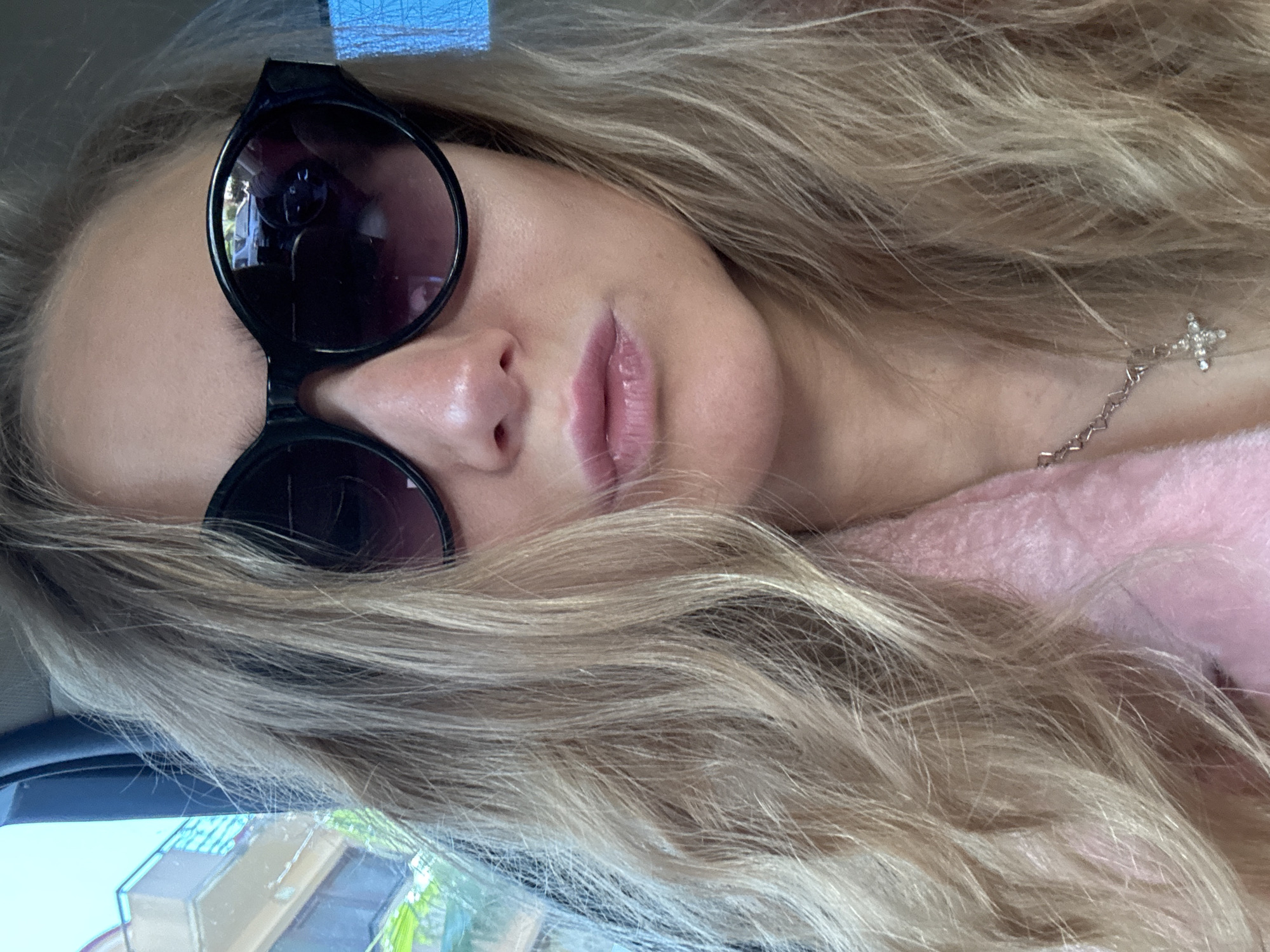 A woman with blonde hair is seen lying down indoors, likely in a vehicle, wearing sunglasses and a necklace with a cross pendant. Buildings and vegetation are visible in the reflection of her sunglasses, and there is a pink, soft fabric visible near her neck. The location is unknown.