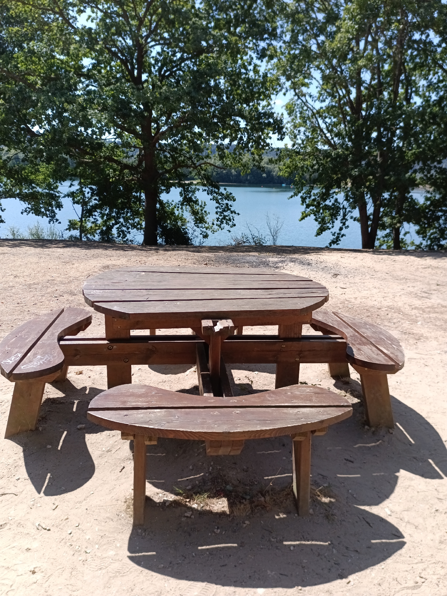 An outdoor picnic table made of wood is positioned on a sandy ground. The table has an oval-shaped tabletop and attached benches. Behind the picnic table, a body of water is visible, likely a lake or river, with trees lining the opposite bank. The sky is clear and blue. The scene appears to be a park or recreational area. This location is in the city of Jumièges, France.