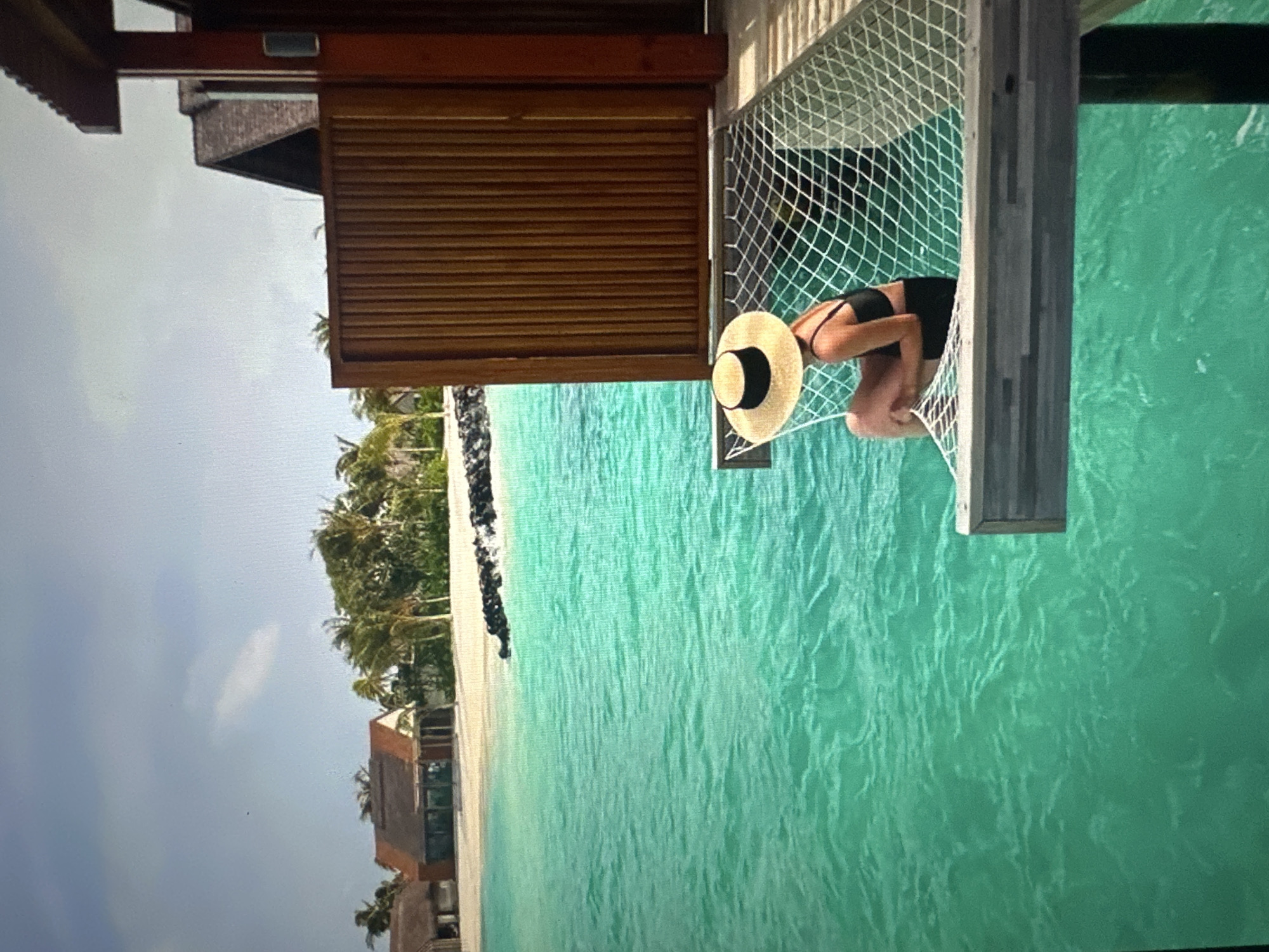 A person wearing a black swimsuit and a straw hat sits in a hammock suspended over turquoise water. The hammock is attached to a wooden deck, part of an overwater bungalow. In the background, a sandy beach is lined with palm trees and other vegetation. Several resort buildings with thatched roofs are visible on the shore. The sky is cloudy with patches of blue.