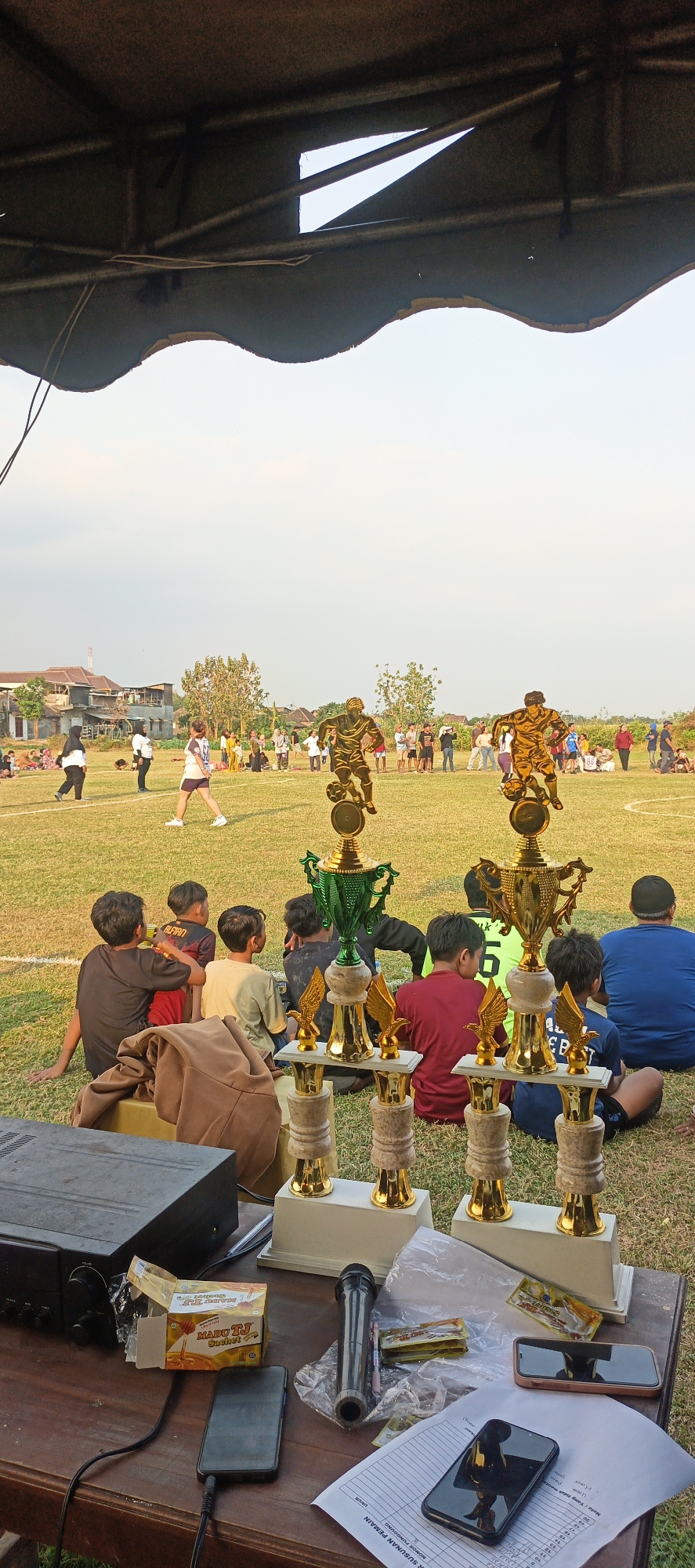 A wide shot captures an outdoor event, likely a sports match, with an audience and trophies on a grassy field under a clear sky. In the foreground, a wooden table displays a black audio amplifier, a box of "Madu TJ" sachets, a microphone, and two smartphones. Several golden trophies with green cups and small eagle figures stand prominently on the grass, some arranged in a row. In the mid-ground, a group of young boys are seated on the grass, facing away from the camera and towards the field. Beyond them, people are gathered on the field, some standing and watching an activity, while others are seated or walking. In the background, houses and trees are visible, with the sky dominating the upper portion of the frame. The overall impression is of a community gathering or a sporting competition.
