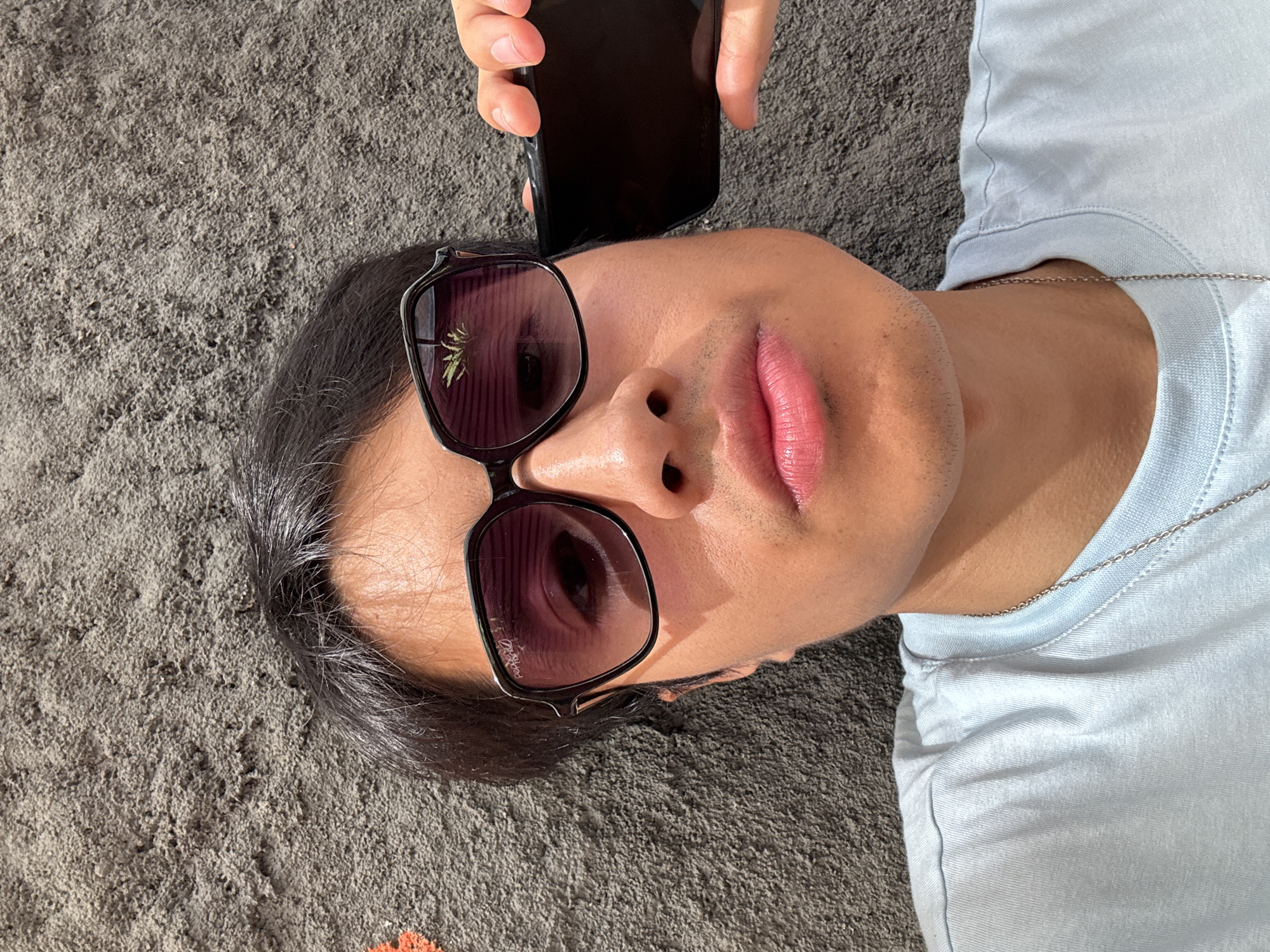A person is lying on a gray surface, possibly sand or a textured fabric, taking a selfie with a black smartphone. The person is wearing sunglasses with a dark frame, reflecting a building or vegetation. They have dark hair, pink lips, and are wearing a light blue t-shirt with a silver chain necklace. The location is likely an urban area, but without clear identifiers from the image, the city of São Paulo, Brazil cannot be definitively confirmed.