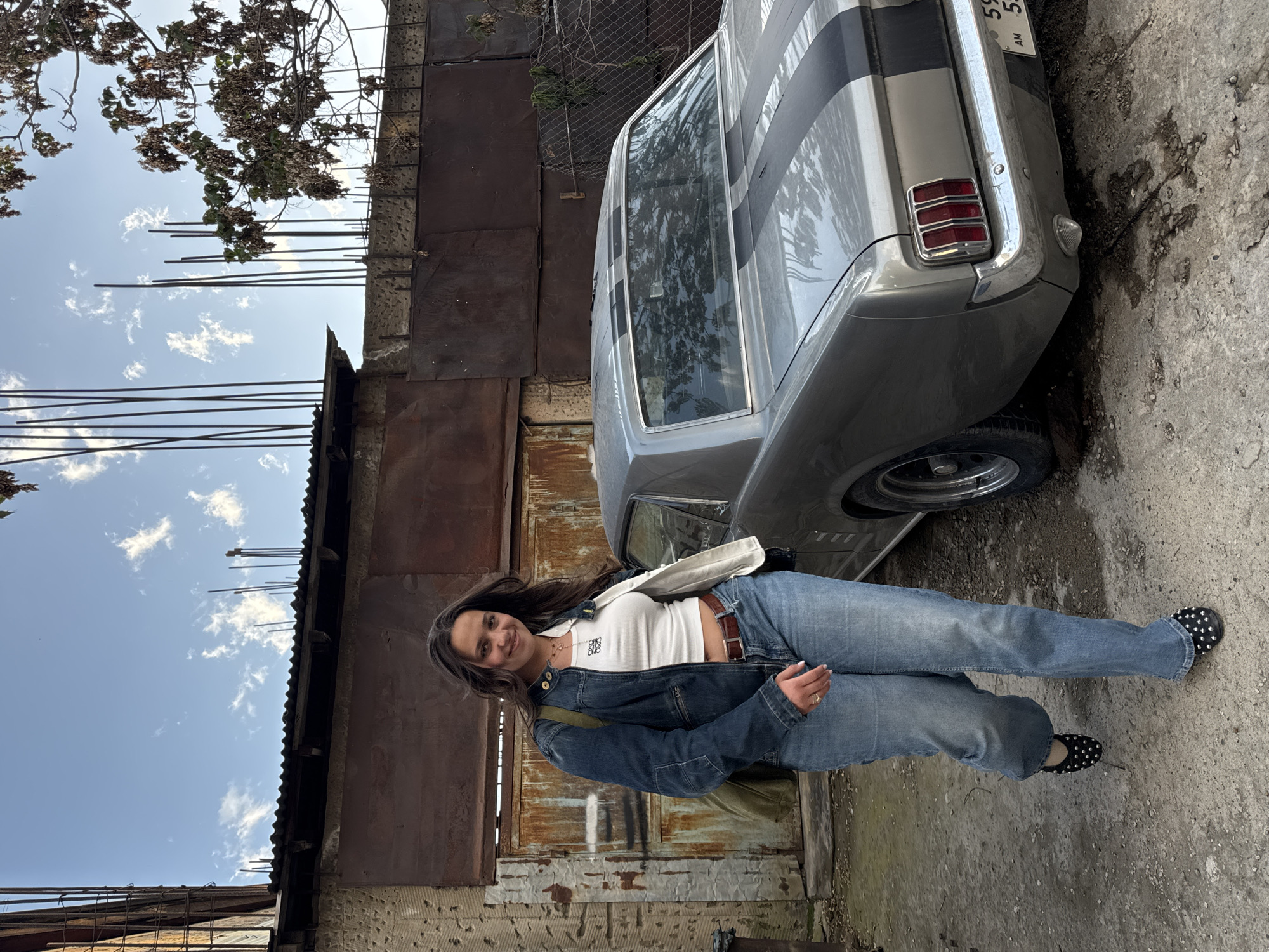 A woman is standing next to a silver and black classic Mustang. She has long dark hair and is wearing a white top, blue denim jacket, and blue jeans. She is smiling and looking at the camera. The car is parked on a concrete surface in front of a rusted metal wall. There is a tree with bare branches in the upper left corner of the image, and a blue sky with white clouds is visible behind it. The location is Yerevan, Armenia, as indicated by the license plate of the car.