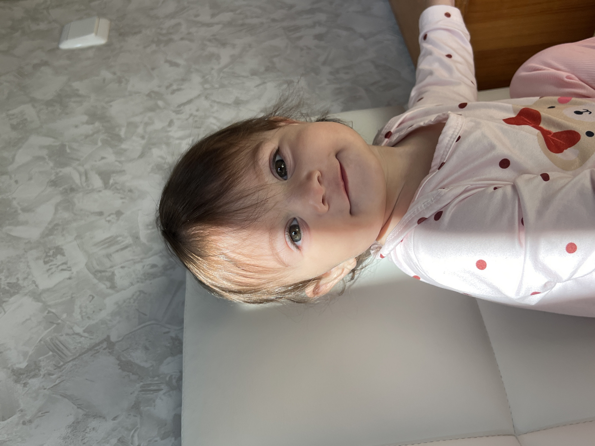 A close-up shot shows a young child wearing pink and white pajamas with red polka dots and a bear motif. The child is lying on a white surface, possibly a bed or couch. The child's head is angled towards the viewer, and they have brown hair, fair skin, and green eyes. The background consists of a grey wall with a textured pattern and a white rectangular object in the upper left corner, and a wooden object in the upper right corner. The location is undetermined.