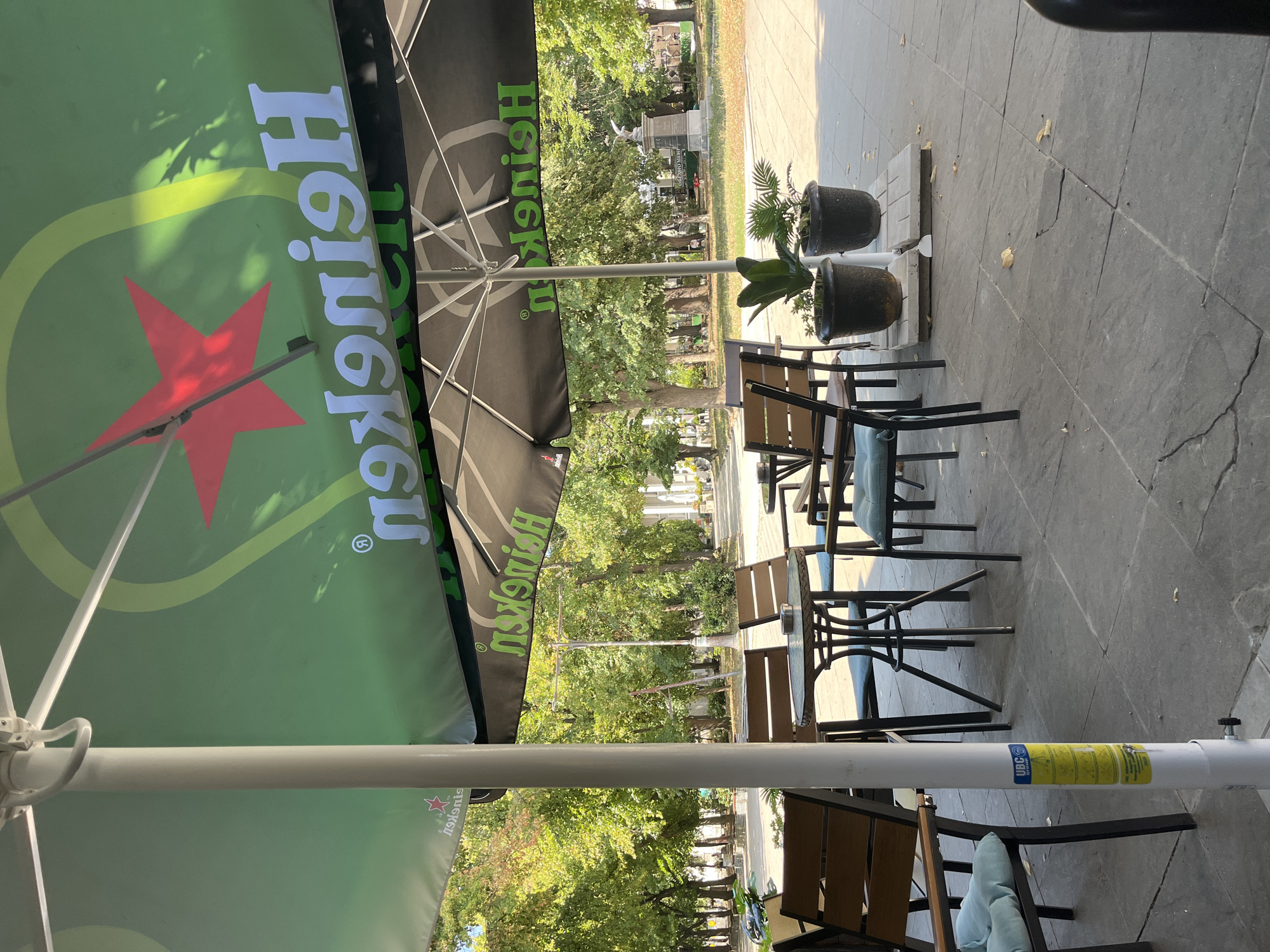 The image depicts an outdoor seating area for a cafe or restaurant in Blagoevgrad, Bulgaria, likely in a park or public square given the trees and statue in the background. The foreground is dominated by a large green and black umbrella with the Heineken logo. Underneath the umbrella, several tables and chairs are arranged. The tables are round with dark metal frames and glass tops. The chairs have dark metal frames with wooden slats for the back and seat, and light blue cushions.
In the background, lush green trees provide shade, and sunlight filters through the leaves, creating dappled patterns on the ground. A statue, likely a monument, is visible in the distance, as well as a street lamp and some buildings. The ground is paved with large, grey square tiles, some of which show signs of wear and cracking. Two potted plants, a larger leafy one and a smaller fern-like one, are placed near the tables, adding a touch of greenery. A yellow sticker with instructions in a foreign language is attached to the white pole supporting the umbrella. No people are visible in the image. The overall scene suggests a peaceful, albeit empty, outdoor dining or relaxation spot on a sunny day.