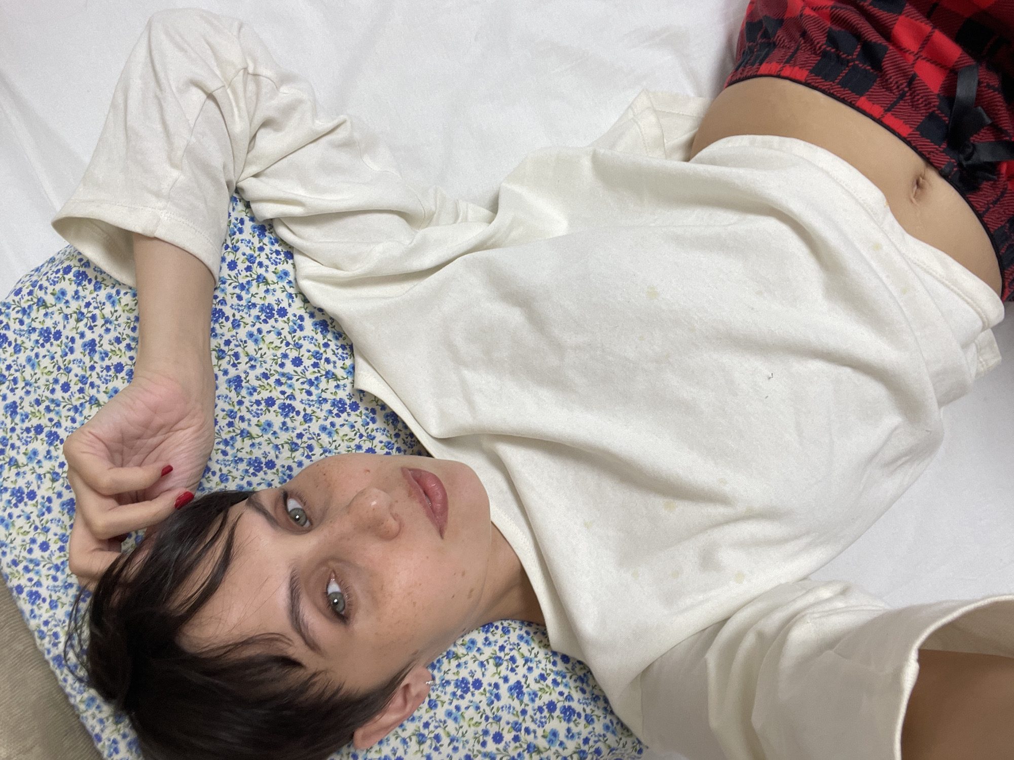A woman is lying on a bed. She is wearing a white shirt and red and black plaid pajama bottoms. Her head is on a pillow with a blue floral pattern. She has brown hair and freckles. Her eyes are open and she is looking at the camera. She is lying on a white sheet. There is no indication of Izhevsk, Russia in the image.