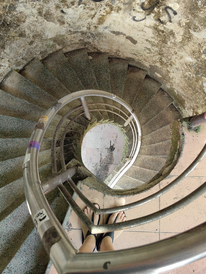 The image displays a high-angle view looking down a weathered spiral staircase located in Thủy Xuân Ward, Vietnam. The staircase features numerous individual stone or concrete steps, each exhibiting a coarse, speckled texture and significant accumulations of green moss and dark grime. The circular walls of the stairwell are constructed from a rough, discolored material, possibly concrete or plaster, showing extensive water damage, peeling, and localized patches of green biological growth. Black graffiti is visible on the upper portion of the wall in the top right quadrant. A multi-barred metal railing, appearing to be stainless steel, spirals downwards, consistent with the staircase's curve. This railing shows signs of wear and contains multiple adhesive stickers, including a distinctive white smiley face icon. At the lowest visible point, the center of the spiral reveals a circular landing paved with light brown square tiles, which appear wet and stained. A pair of bare human feet, wearing black flip-flop sandals, are positioned on the uppermost visible landing, directly beneath the camera's perspective, indicating the presence of an individual observing the descent.